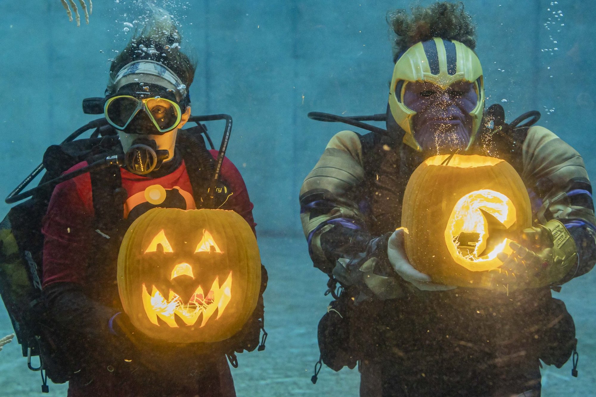 Two costumed scuba divers hold glowing carved pumpkins underwater in an aquarium pool.