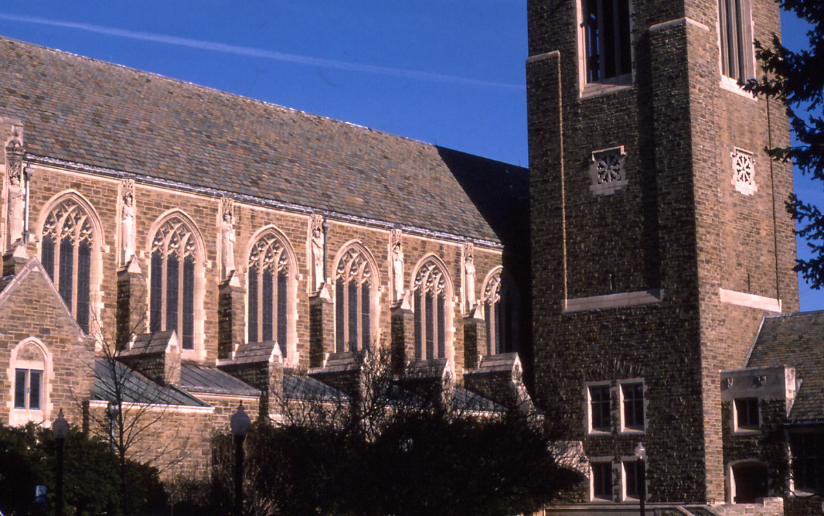 Gothic stone church exterior with large arched windows and a square tower amid trees.