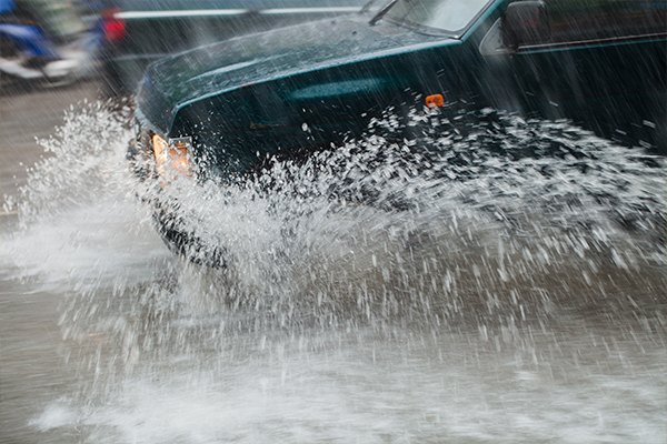 A vehicle drives through deep standing floodwater on a road in heavy rain.