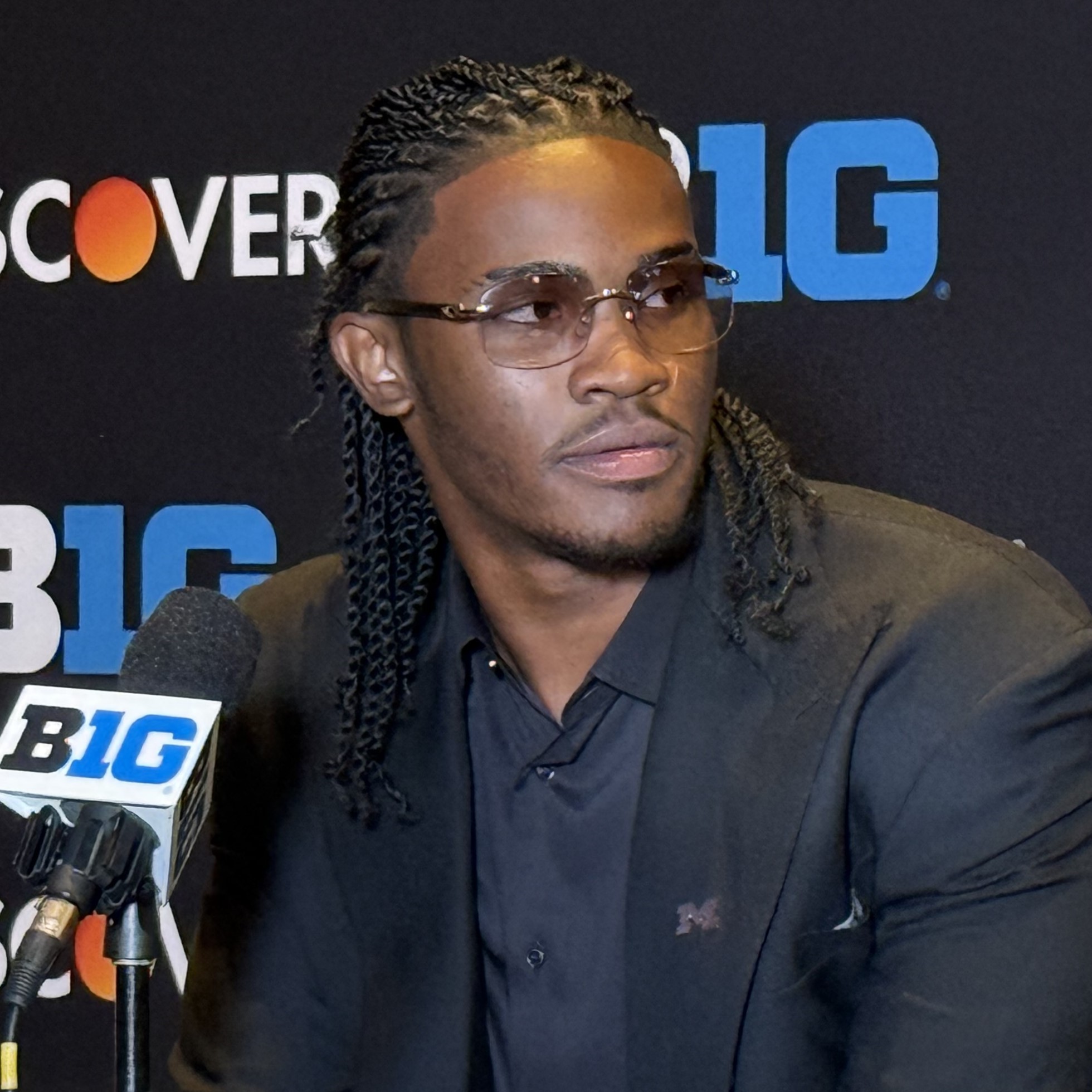 Young man with braided hair in a black blazer at a Big Ten media podium.