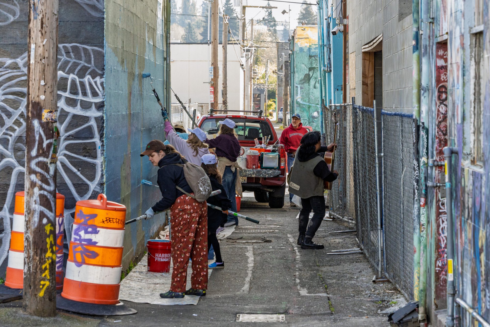 Several volunteers with paint rollers work along a narrow urban alley with graffiti-covered walls.