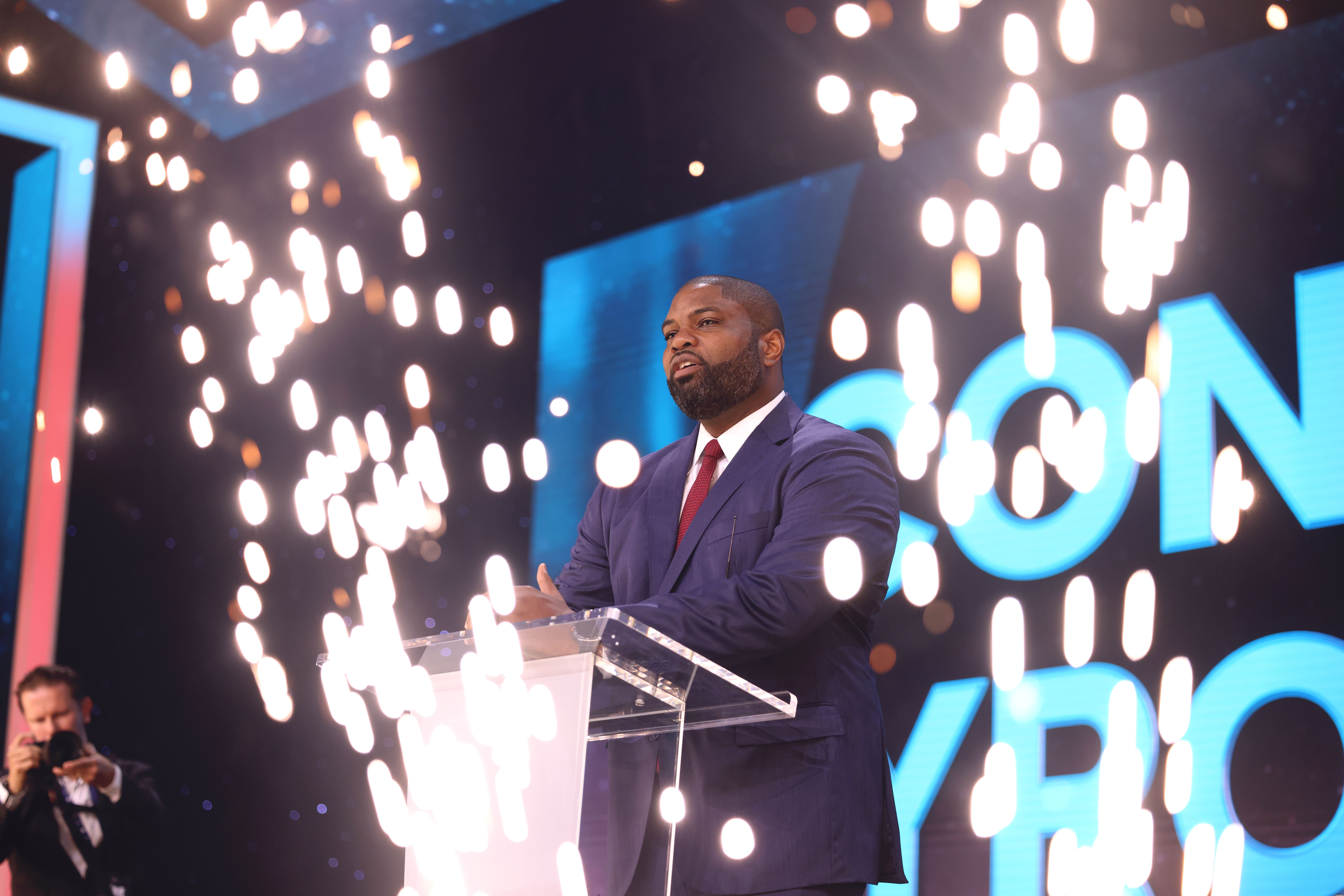 Man in navy suit and red tie speaking at a clear podium on a brightly lit stage.