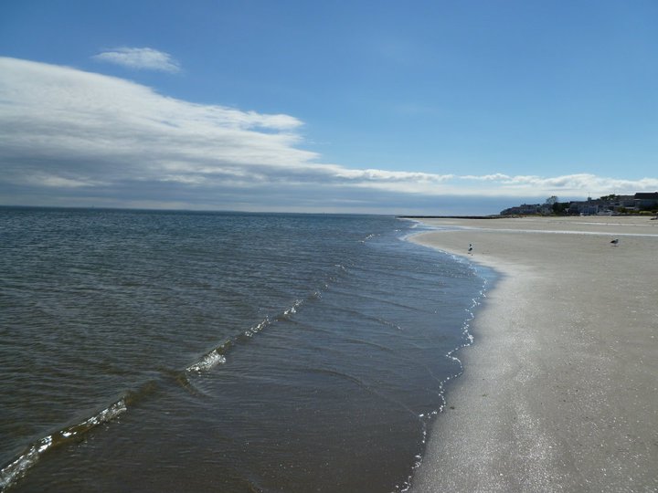 Wide sandy beach along Long Island Sound under a partly cloudy sky with calm water.