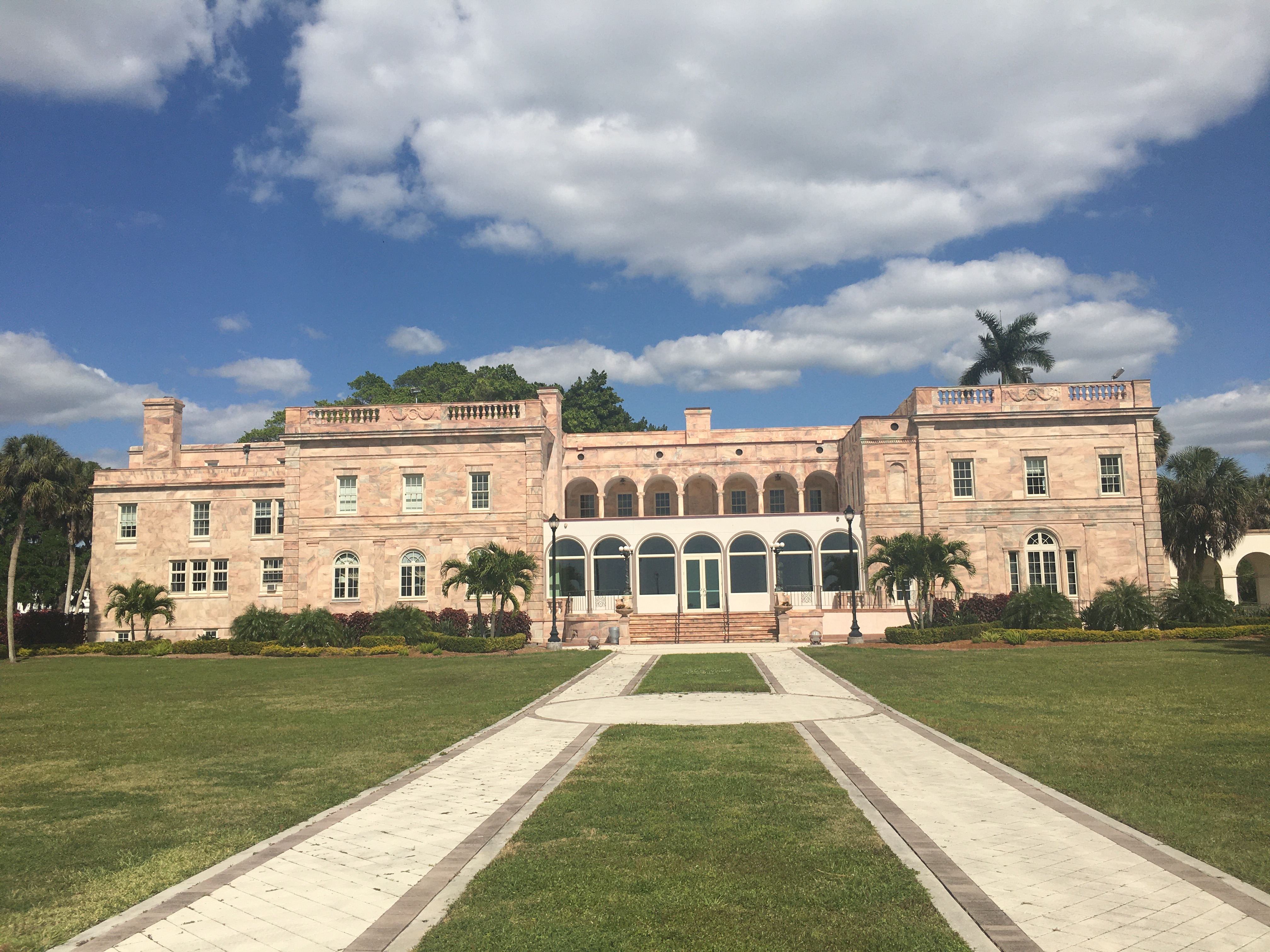 Two-story pink marble building with arched colonnades and palm trees on a sunny day.