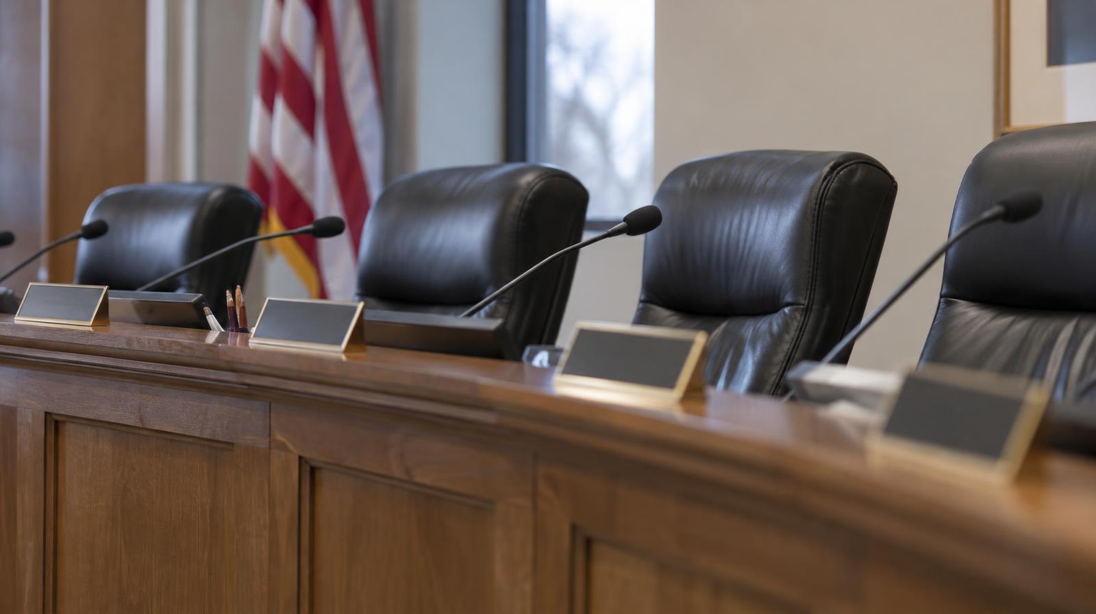 Empty chairs behind a long wooden dais with name placards in a meeting room.