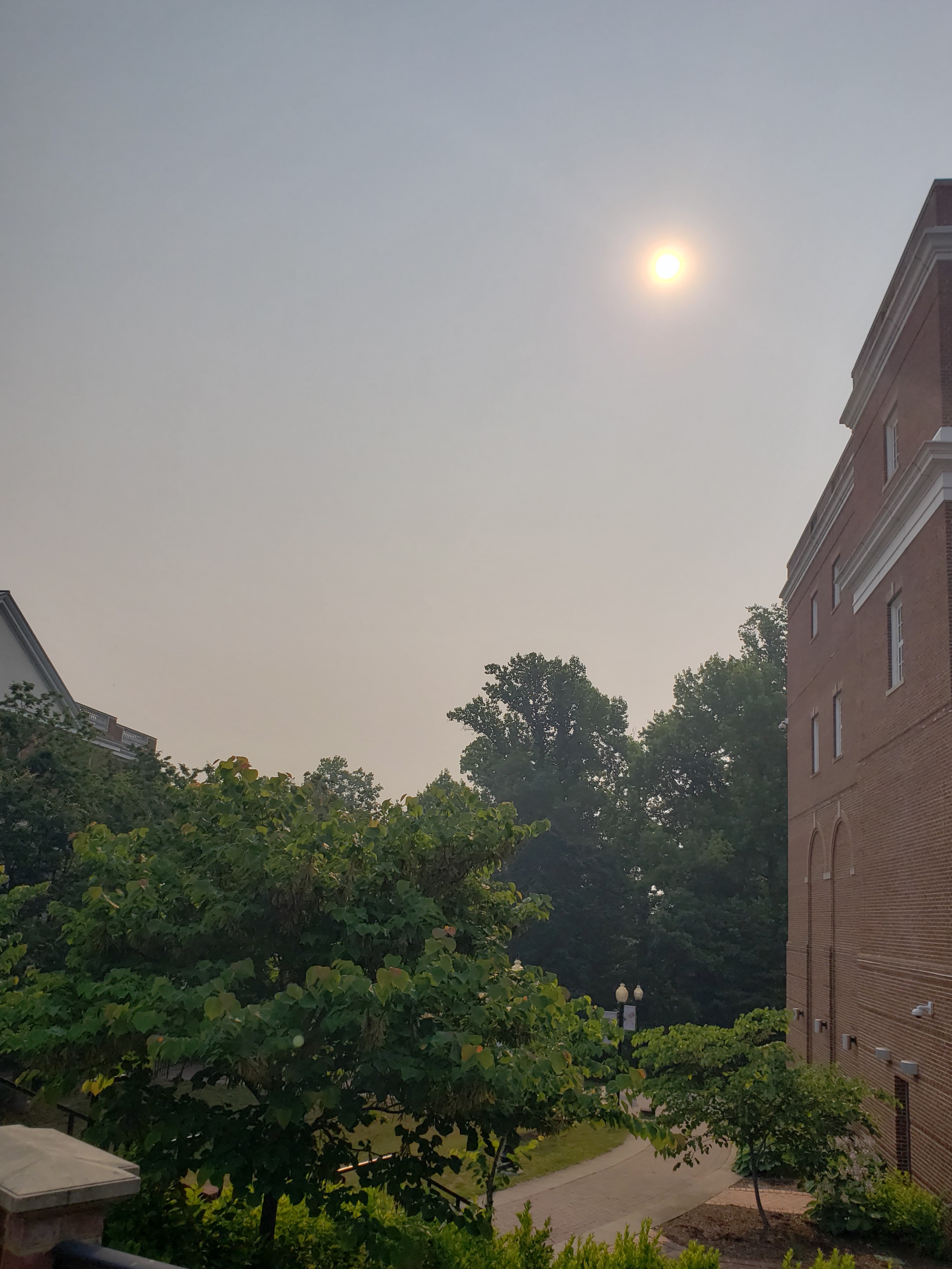 Sun barely visible through a gray-orange hazy sky with brick buildings and trees below.