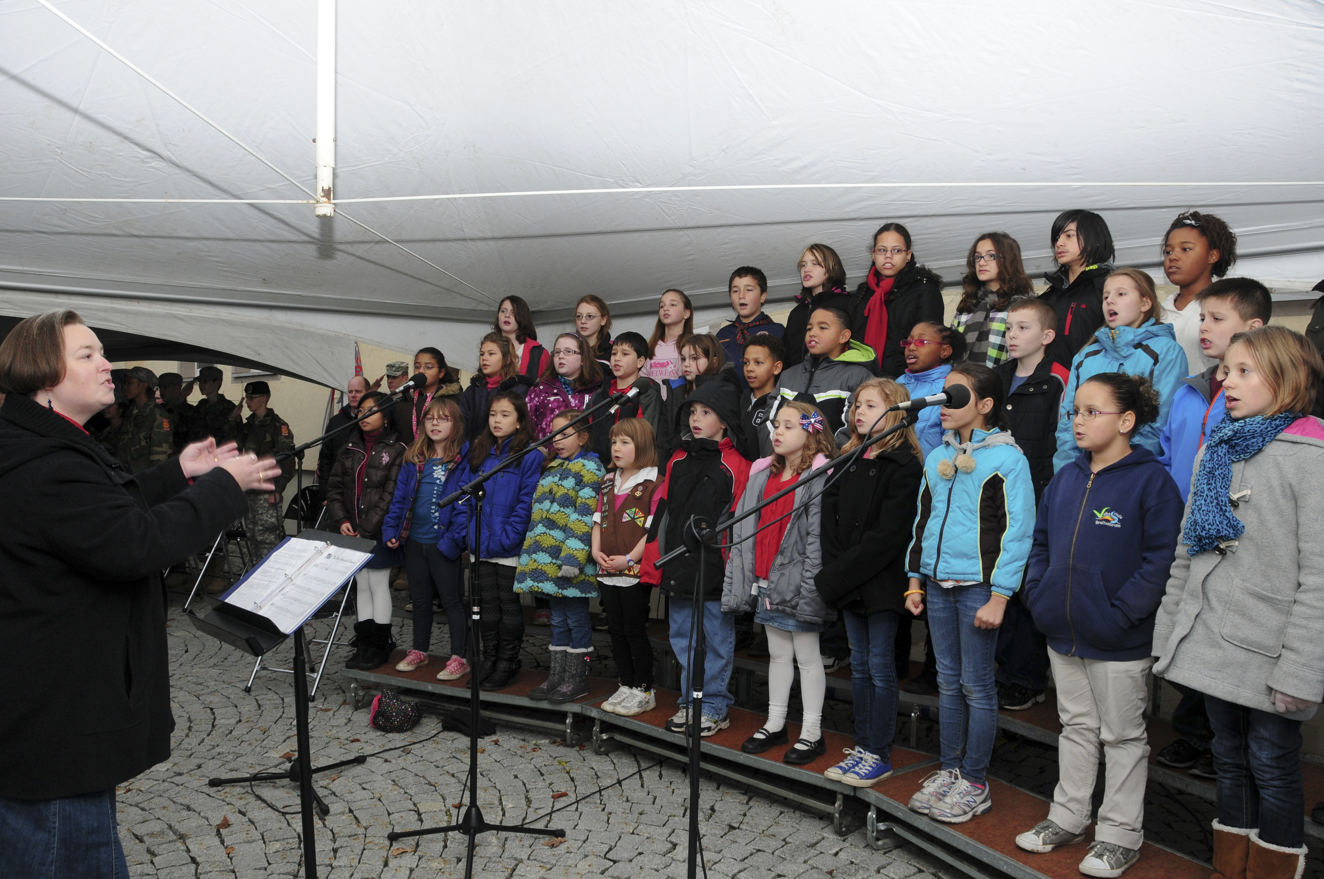 Rows of young children singing on risers at an outdoor Veterans Day ceremony.