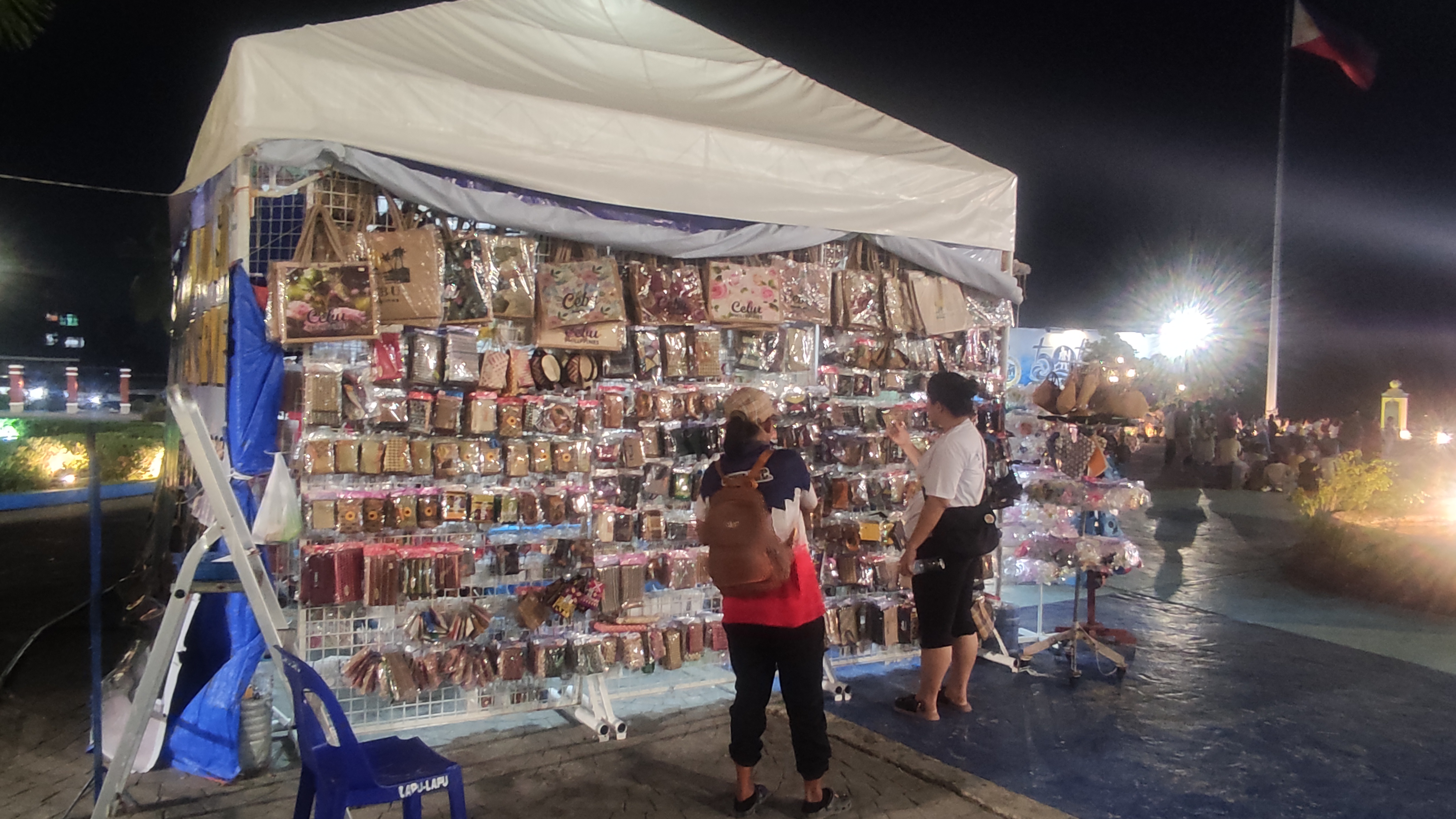 Two shoppers browse a vendor stall packed with bags and accessories under a white tent canopy at night.