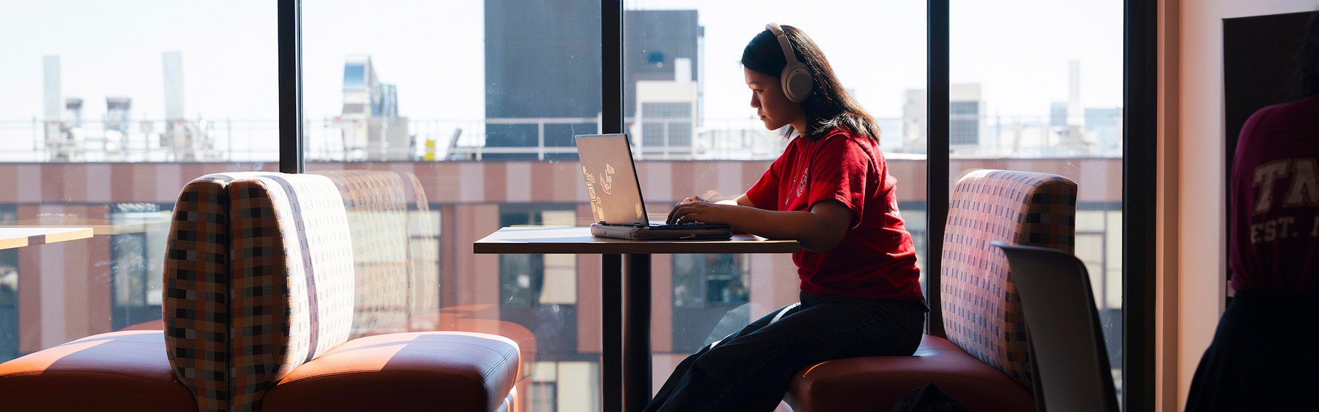 Student in red shirt and headphones works on a laptop near large windows.