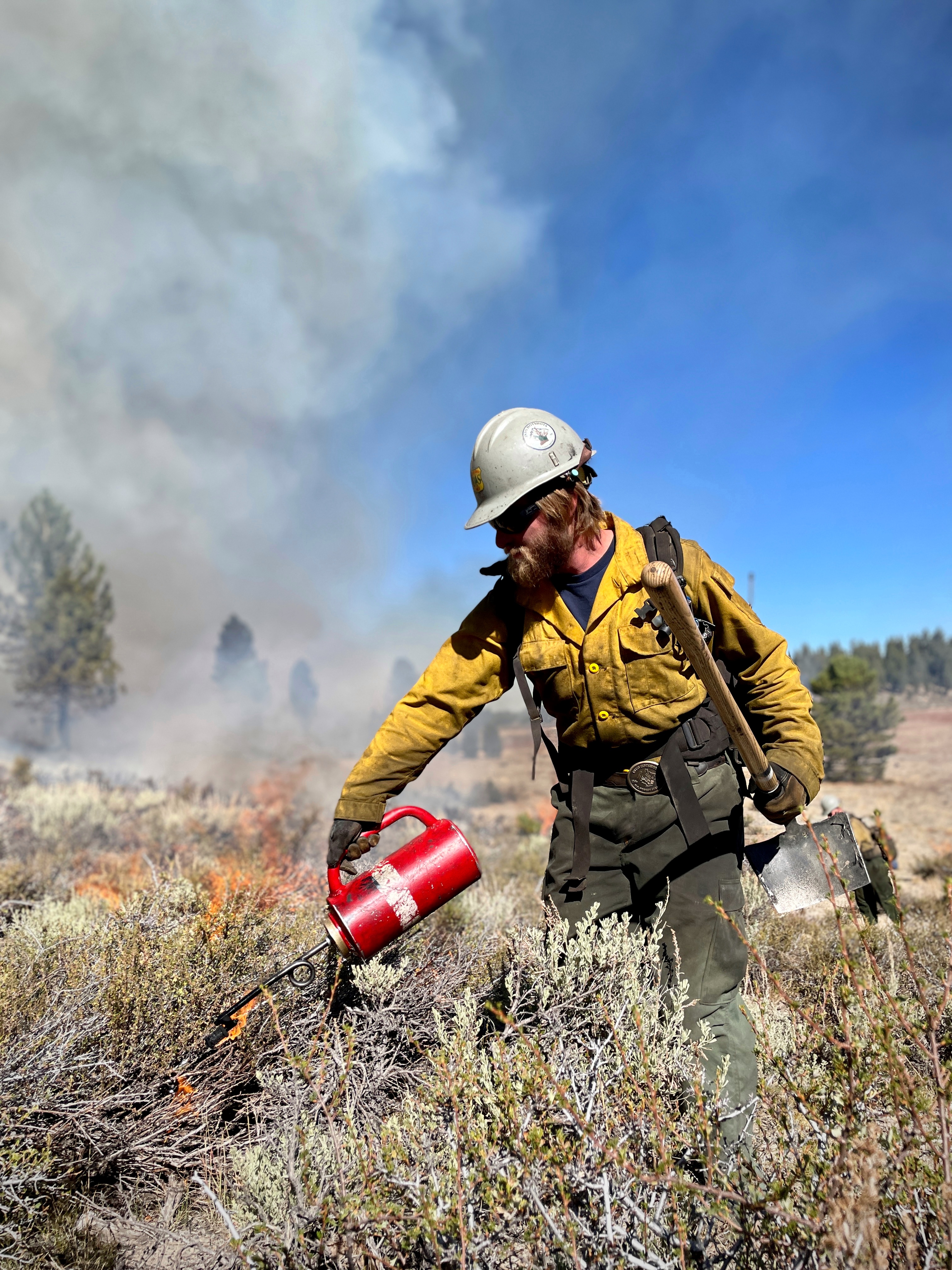 Firefighter in yellow gear holds a drip torch as large smoke plumes rise behind.