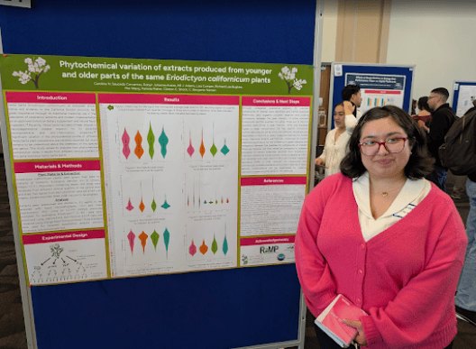 A woman in a pink cardigan stands next to a large scientific research poster at an academic event.