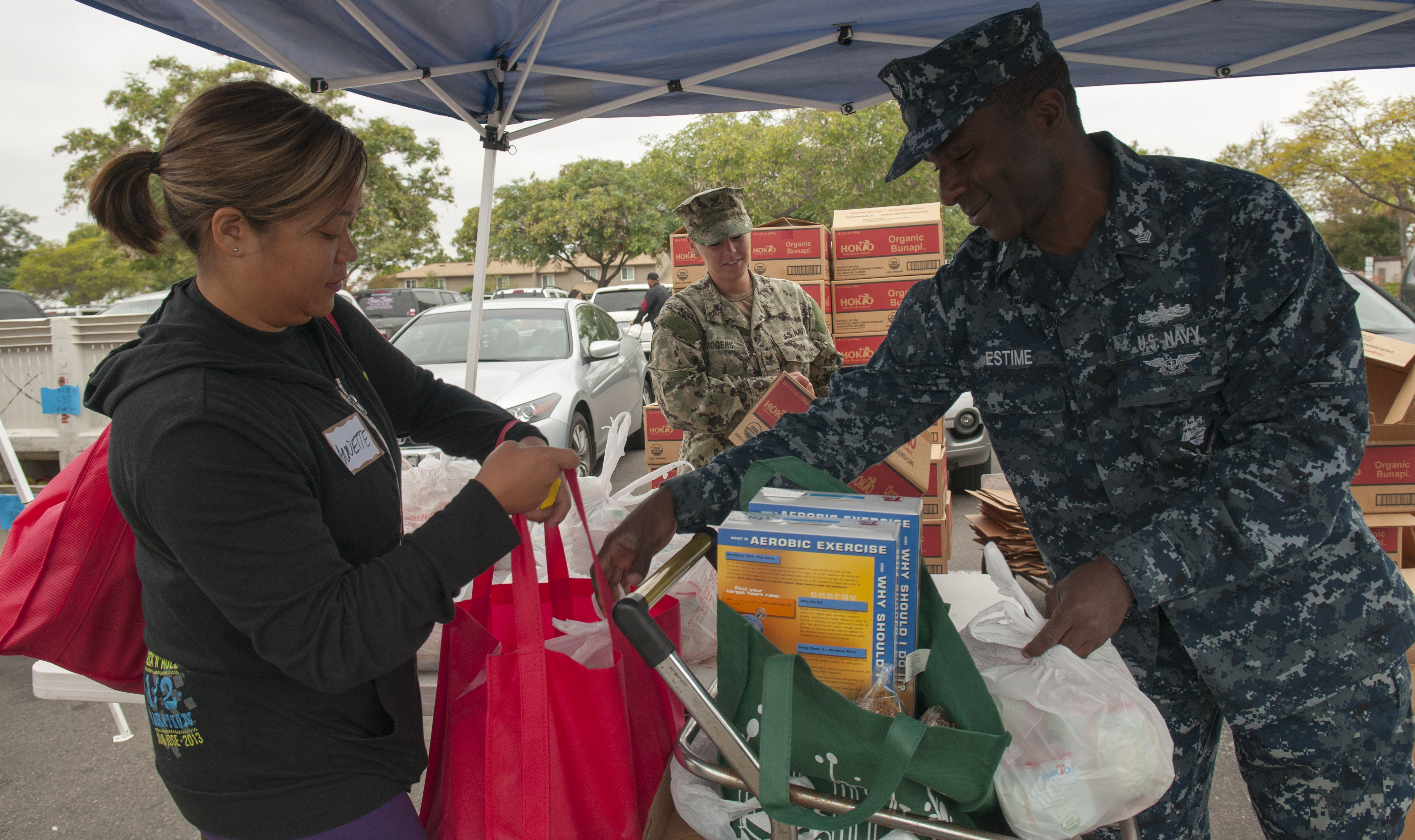 A volunteer hands grocery bags to a woman at an outdoor food distribution tent.