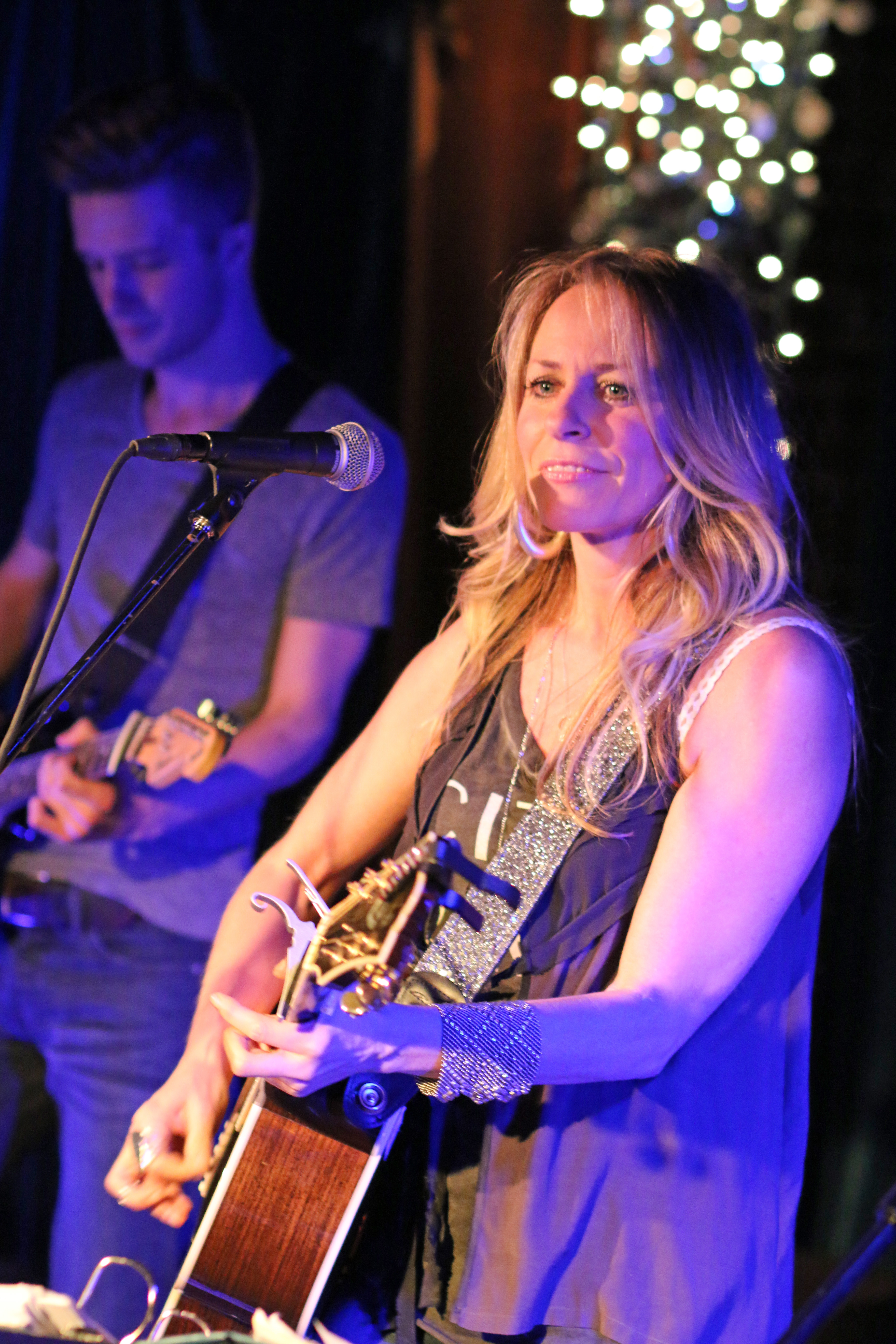 Blonde woman playing acoustic guitar at a microphone on a dimly lit stage.