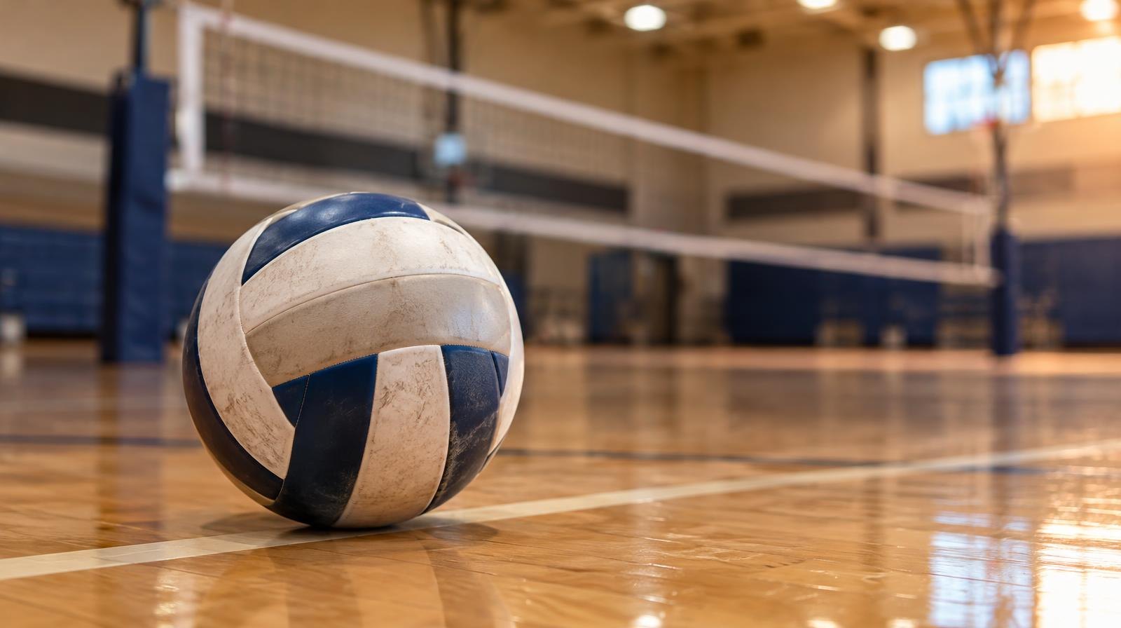 A volleyball resting on a hardwood gym floor near a volleyball net.
