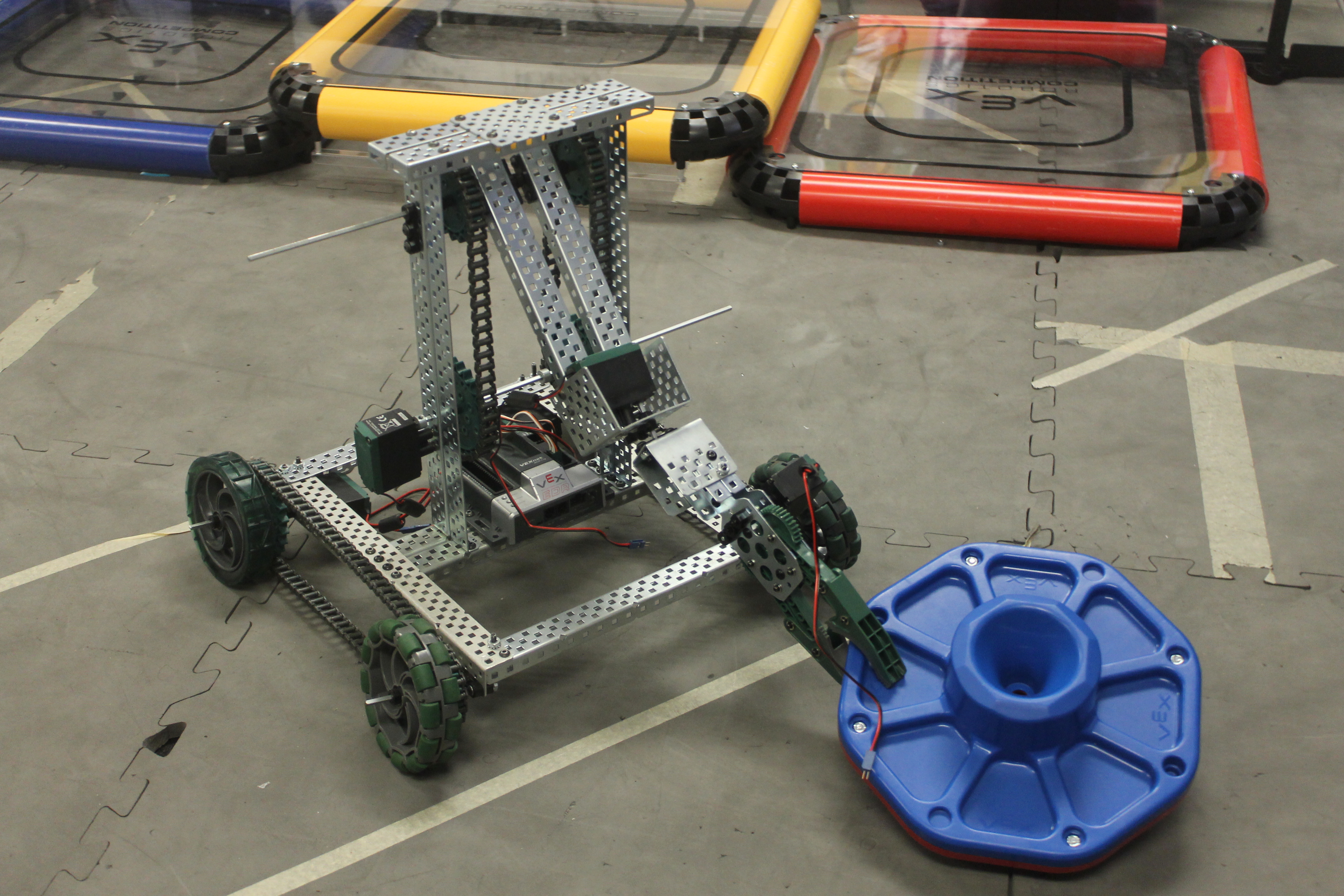 A metal student-built robot with green wheels on a robotics competition field mat.