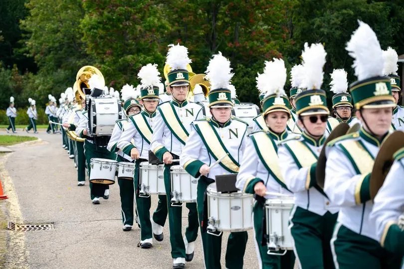 Marching band members in green and white uniforms march down an outdoor path.