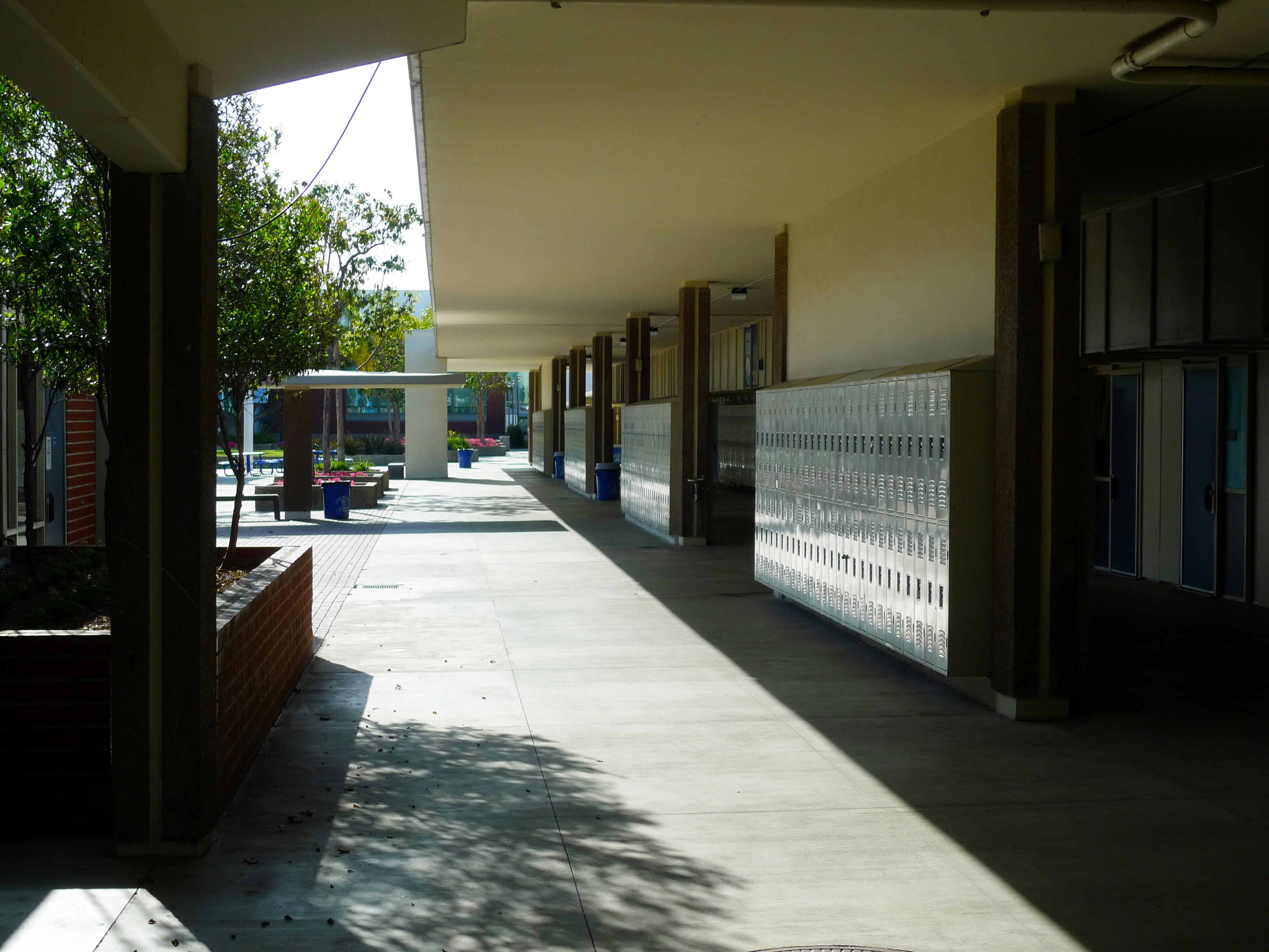 Covered outdoor school hallway with metal lockers lining one side and a tree-filled courtyard beyond.