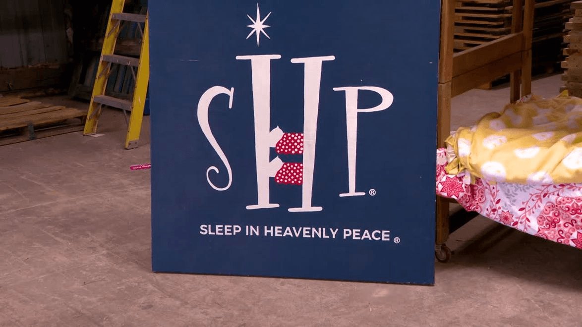 Navy blue Sleep in Heavenly Peace sign leaning against a warehouse wall with lumber in the background.