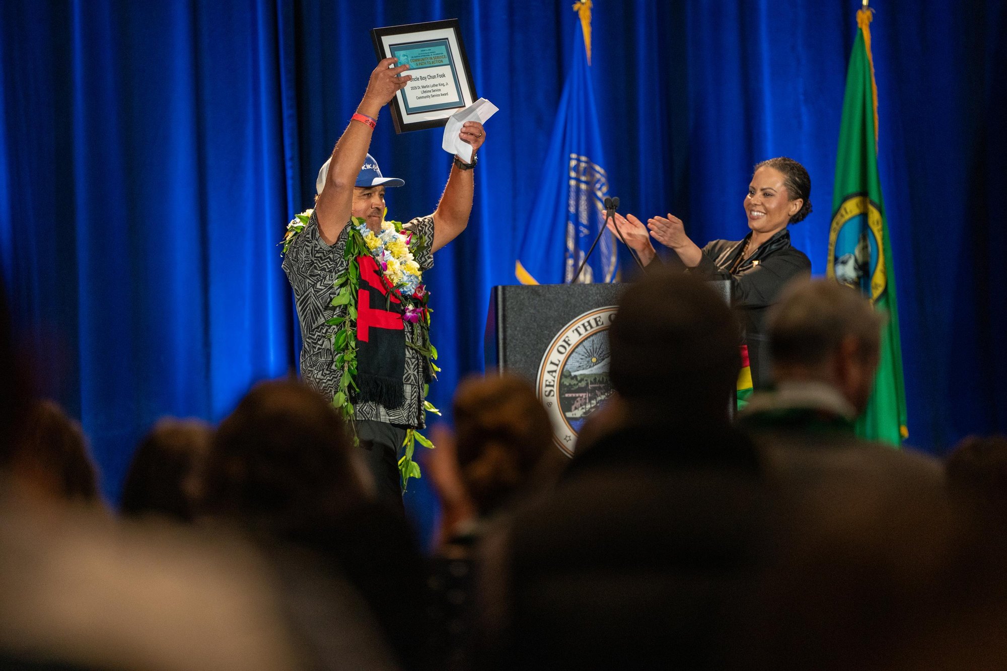 A person in a lei raises a framed award overhead on a stage while another person applauds at a podium.