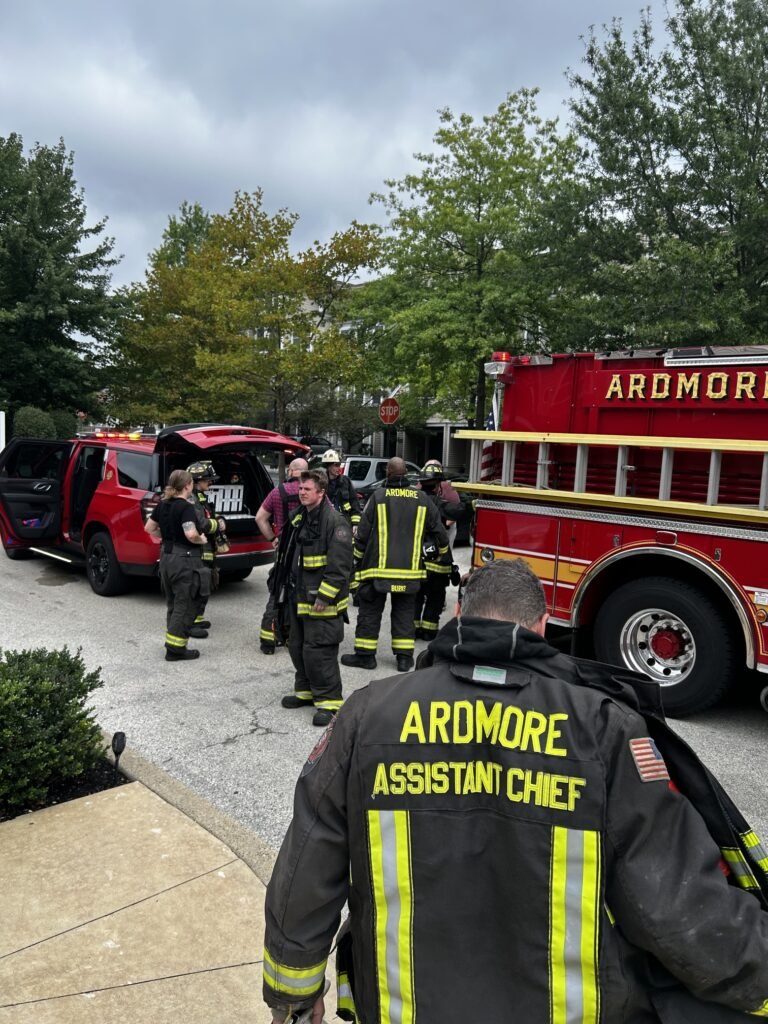 Firefighters in turnout gear stand near a red fire engine marked Ardmore on an overcast day.