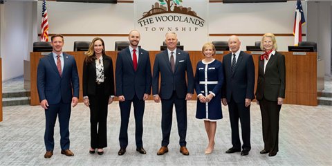 Seven people in formal attire standing in front of a government boardroom dais.