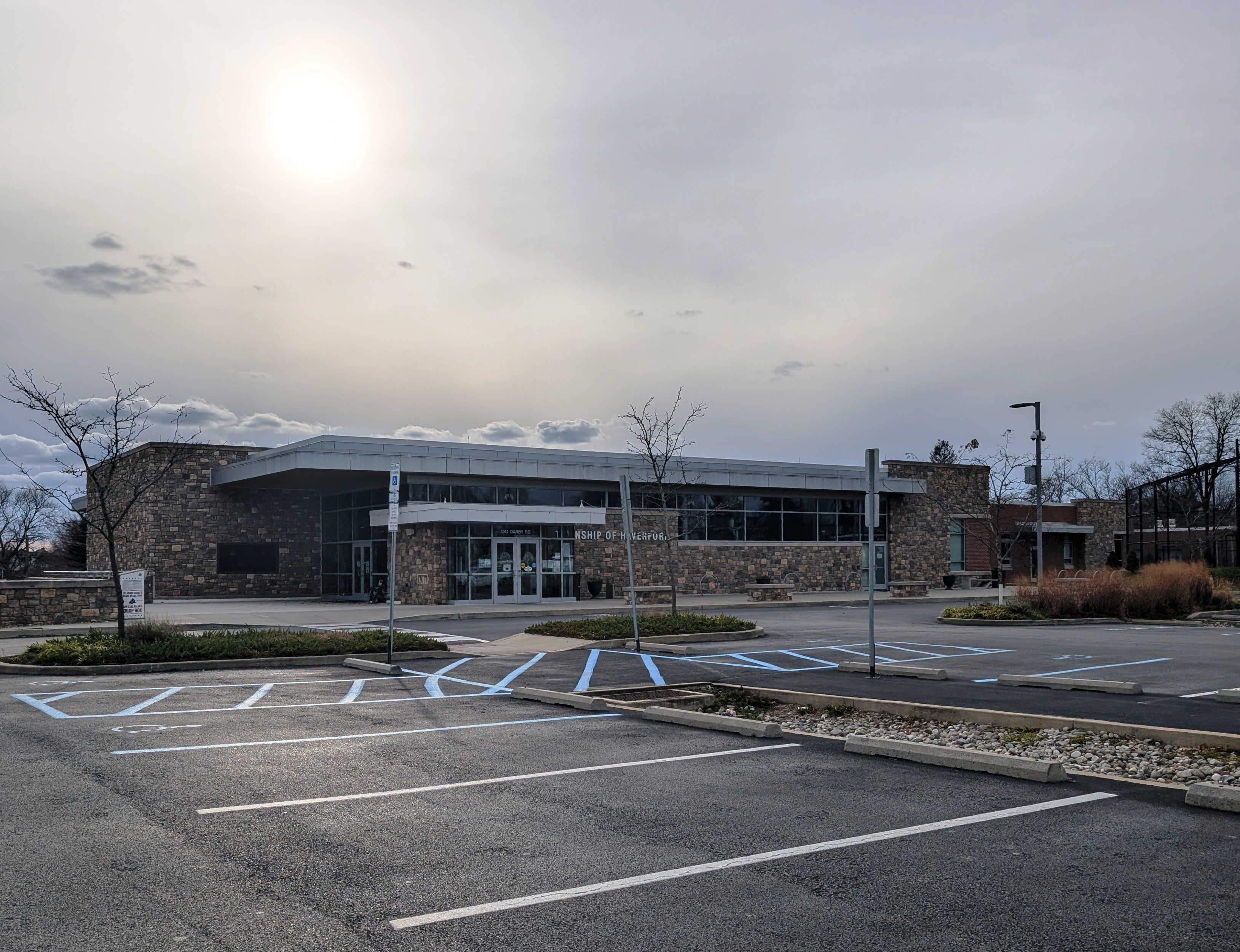 Stone and glass municipal building with parking lot and bare trees under overcast sky.