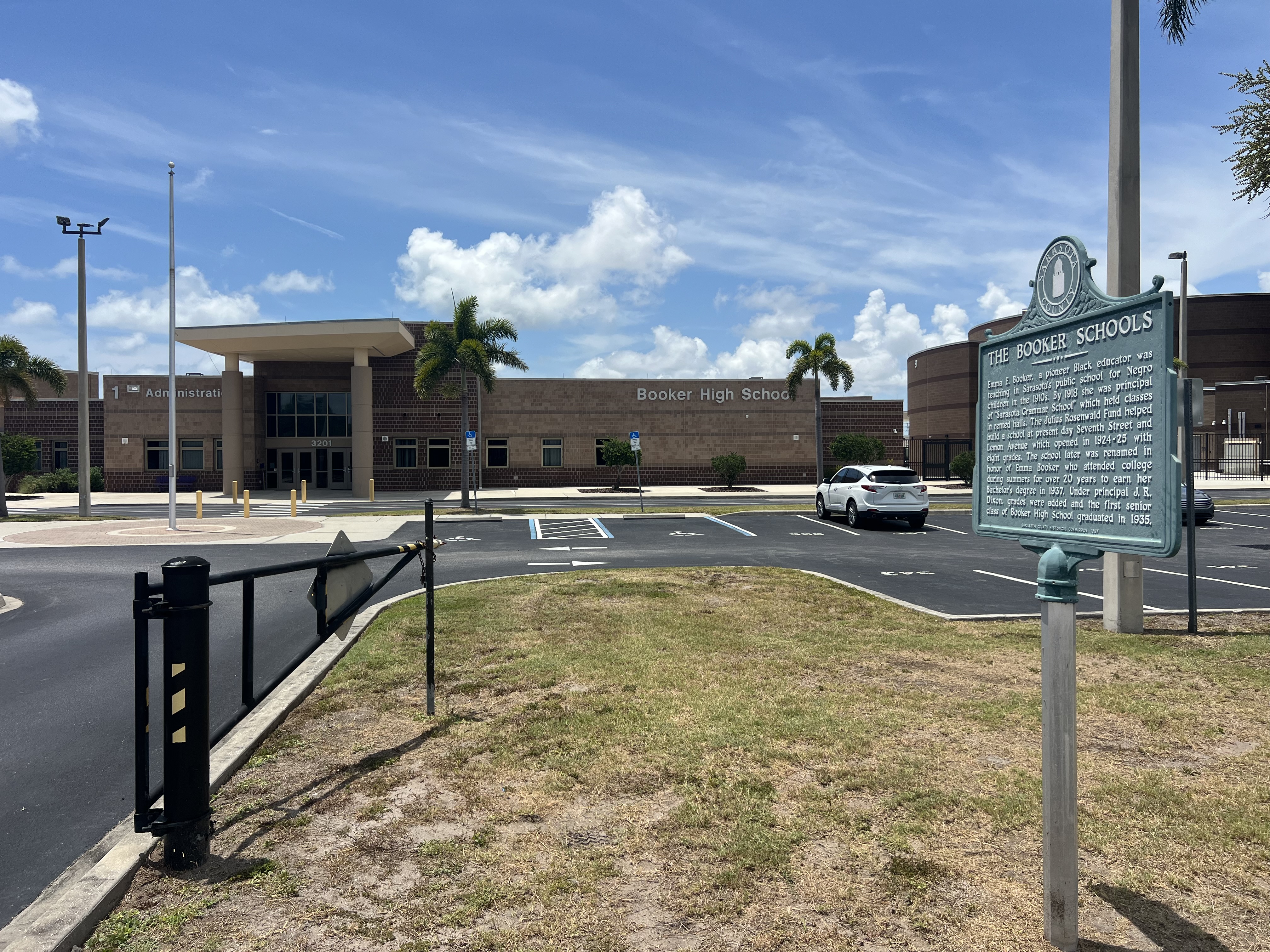 Brick school building with a green historical marker in the foreground and palm trees.