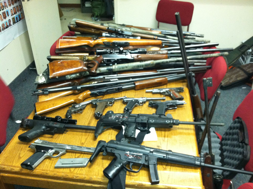 Rows of confiscated rifles, shotguns, and handguns laid out on a wooden table.