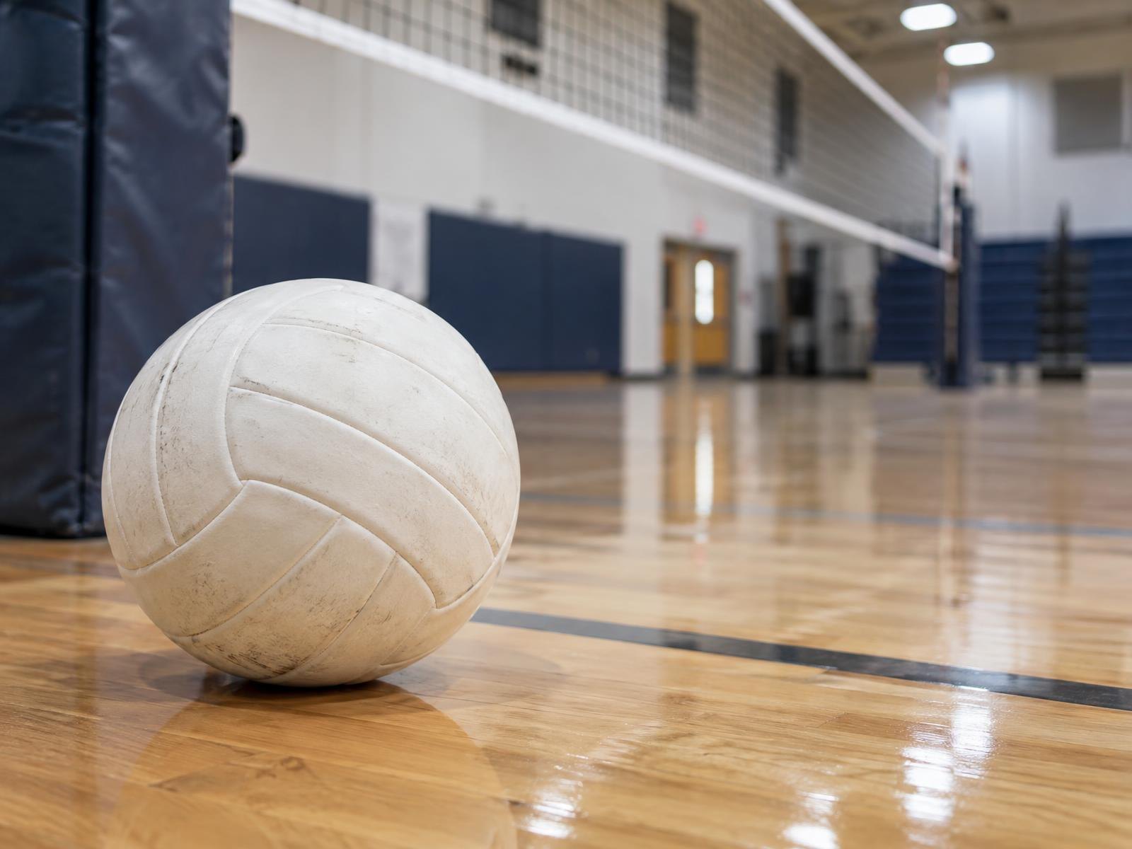 A white volleyball resting on a hardwood gymnasium floor near a net post.