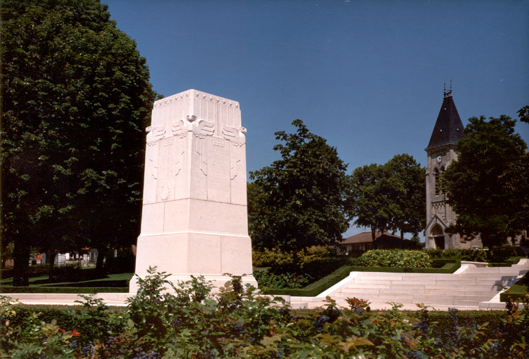 Large white stone memorial monument with relief carvings surrounded by trees and flowers in a park.