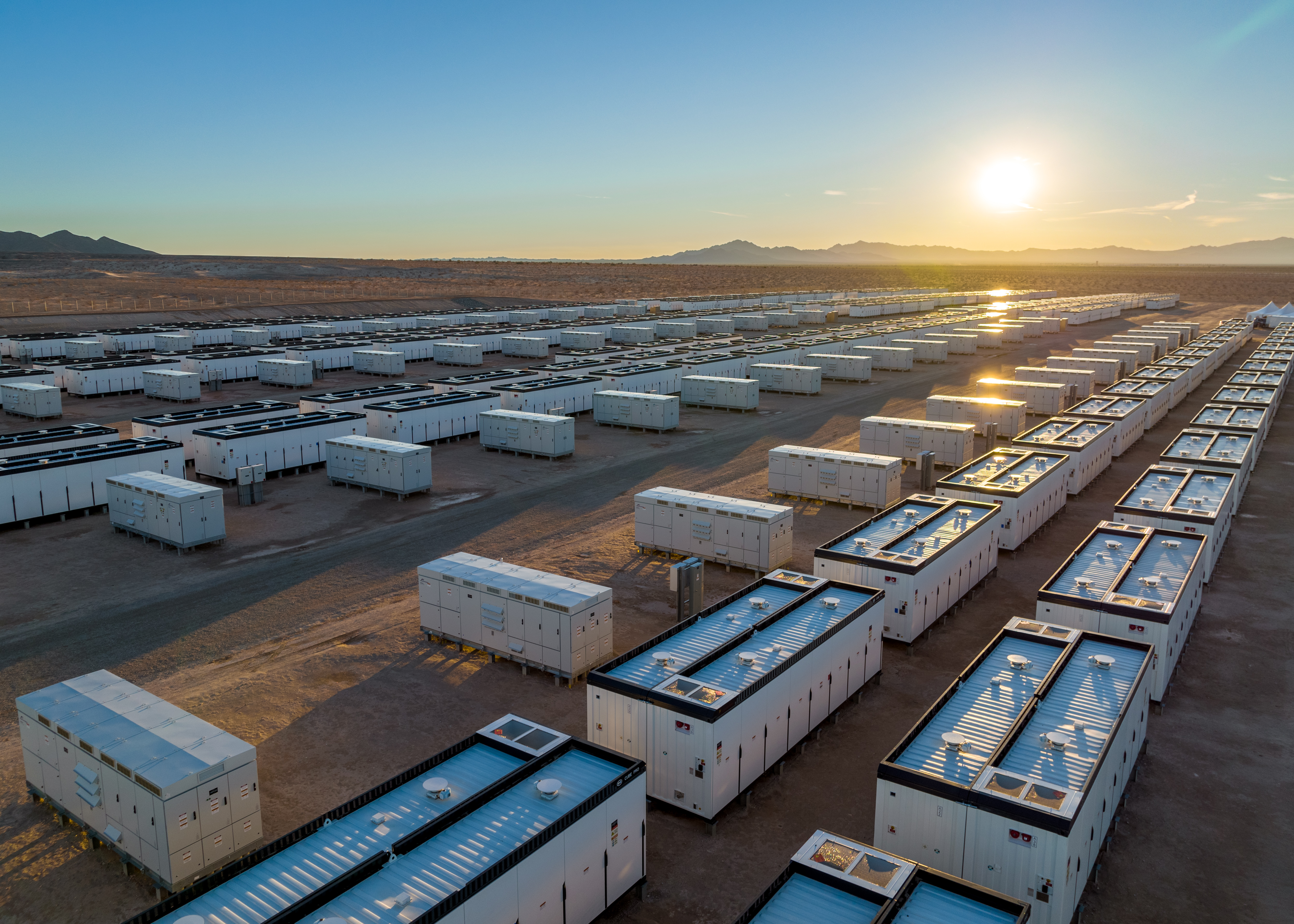 Aerial view of rows of white modular battery storage containers in a desert landscape at sunset.