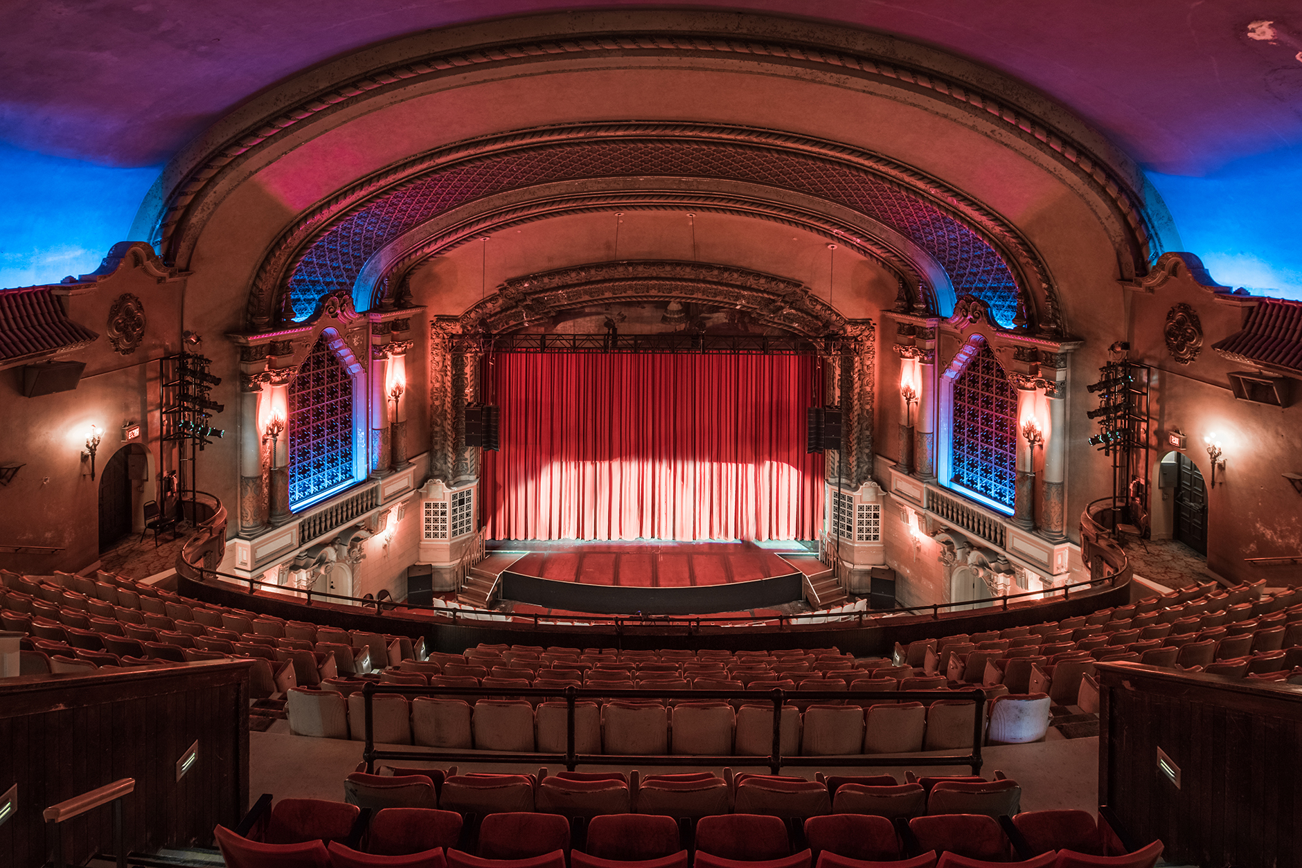 Grand theater auditorium with red stage curtain, tiered red seats, and ornate ceiling lighting.