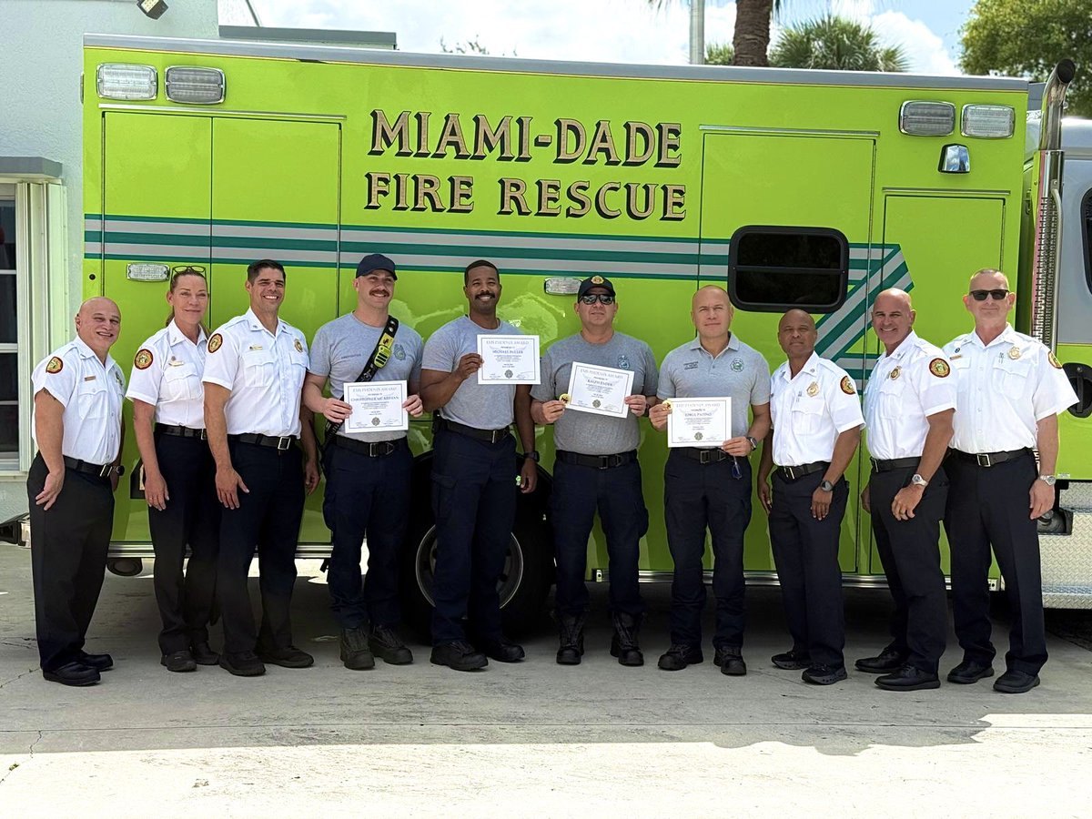Group of fire rescue personnel in uniform posing in front of a lime-green fire rescue vehicle, several holding certificates.