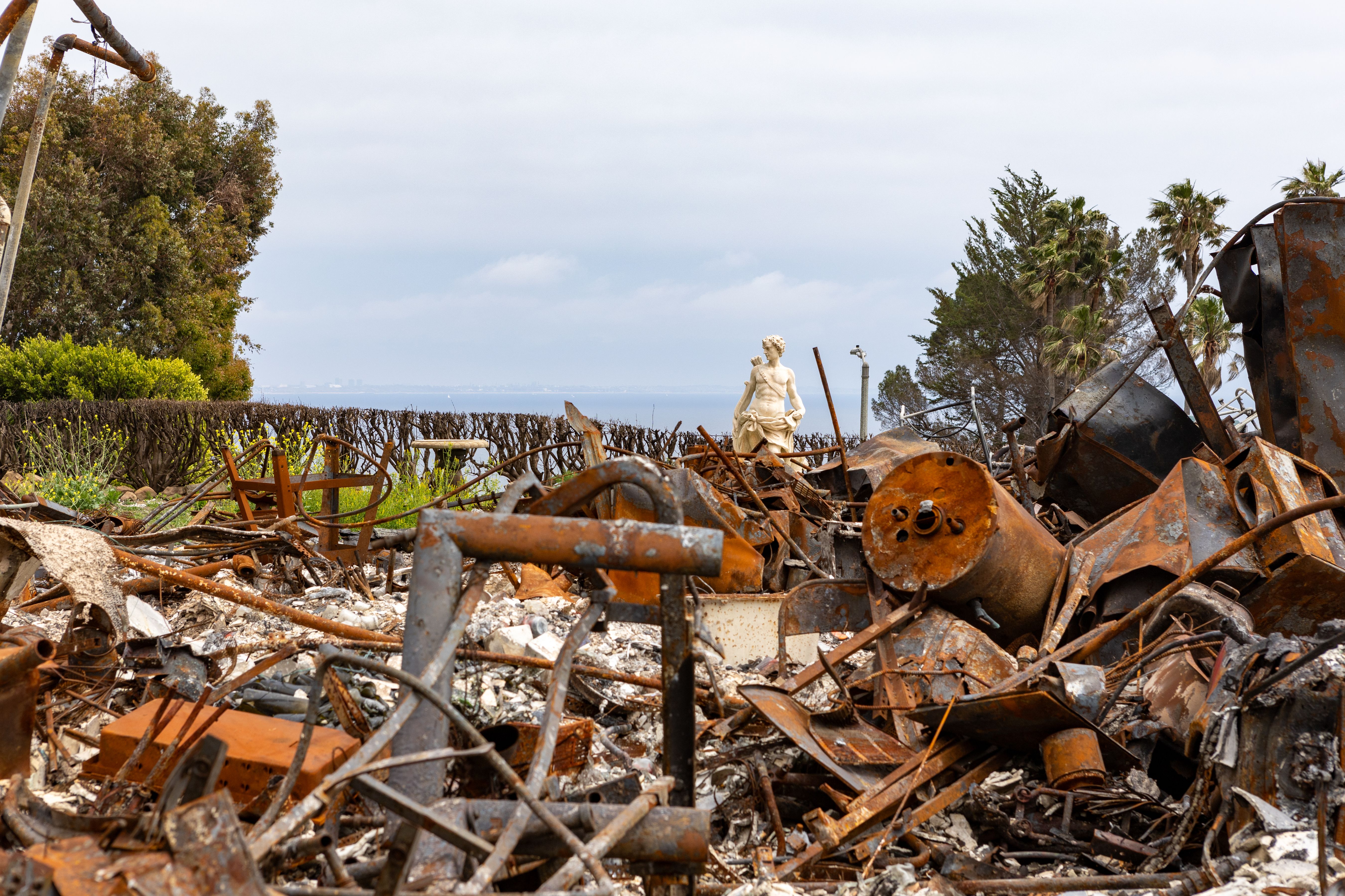 Charred ruins and twisted metal debris from a fire-destroyed home, with trees and water visible in the background.