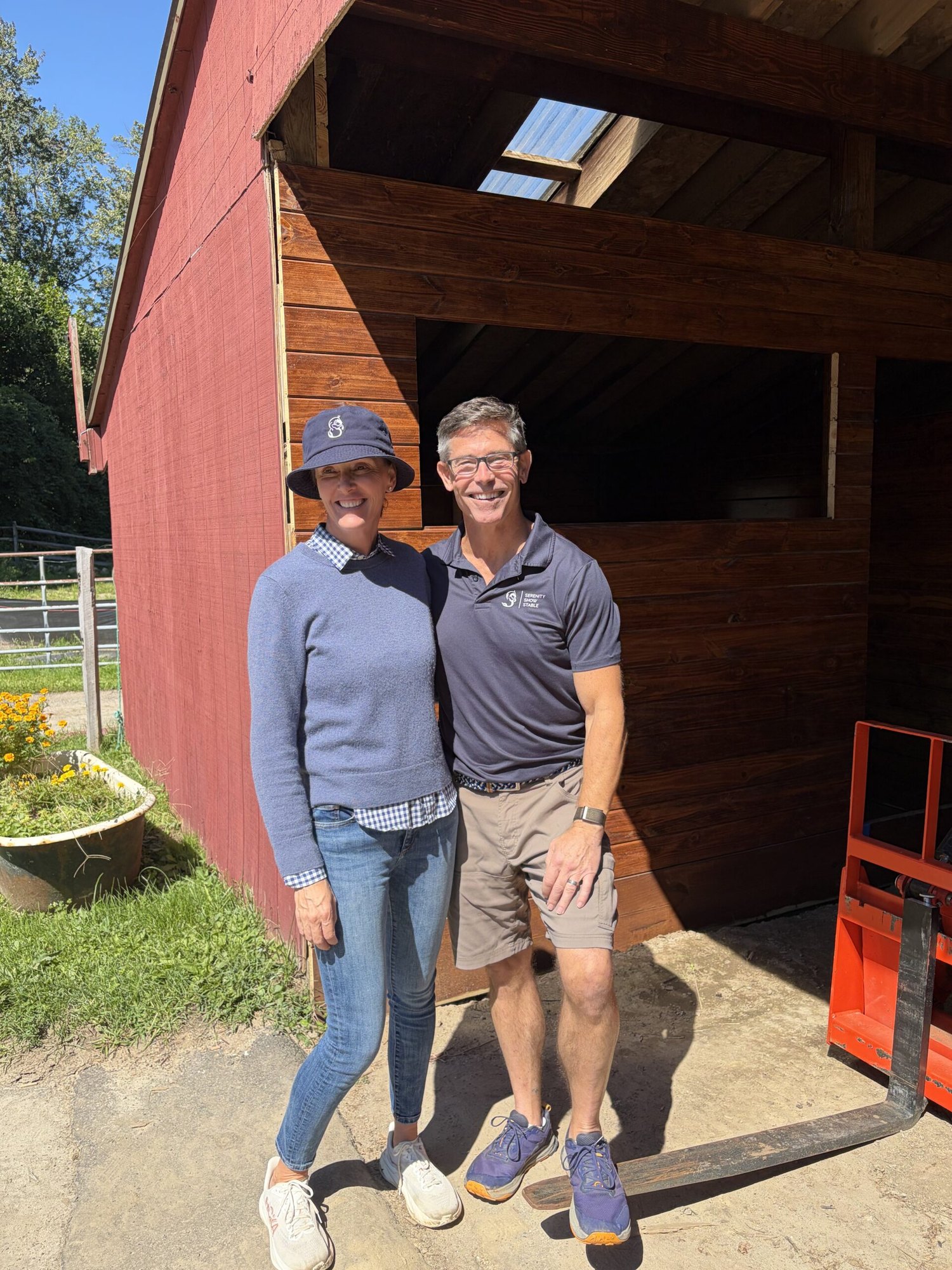A woman and man stand smiling in front of a red wooden barn on a sunny day.