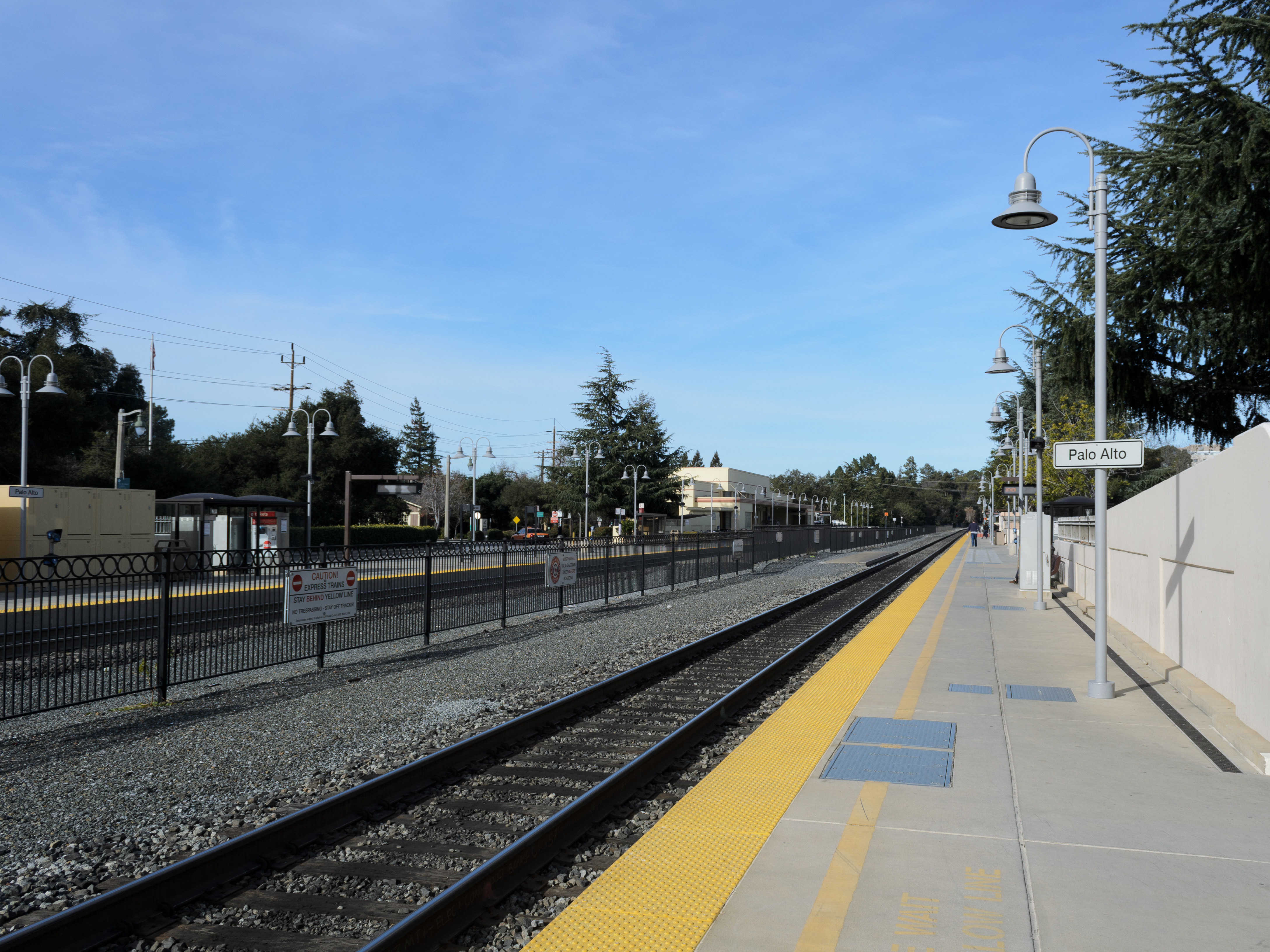 Empty Caltrain station platform with railroad tracks, a station sign, and lamp posts under a blue sky.