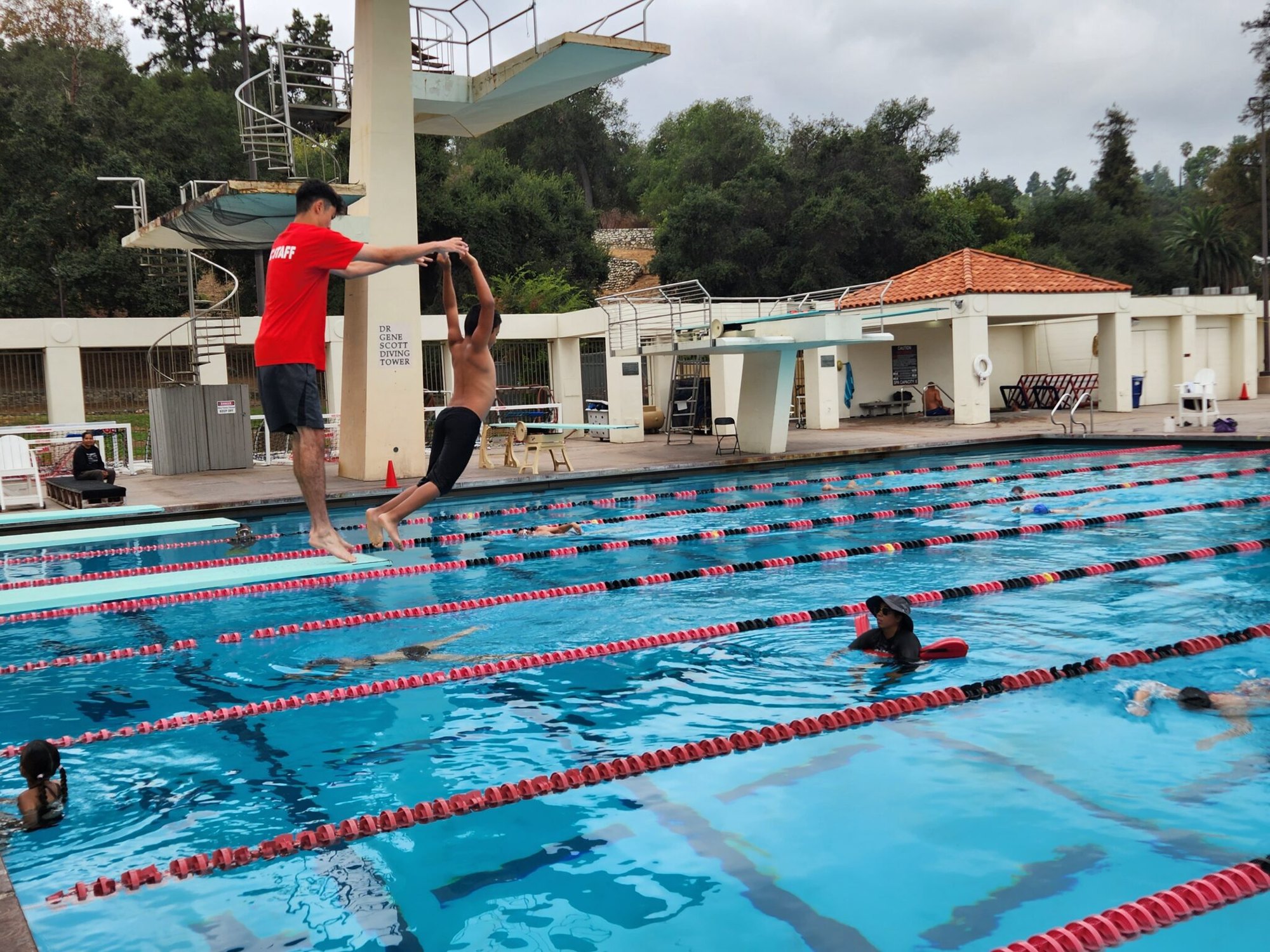 An instructor in a red shirt assists a child at the edge of an outdoor lap pool with lane dividers and swimmers in the water.