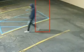 Blurry surveillance image of a hooded person walking across a parking lot beside a red tracking box.