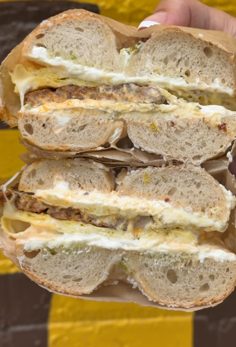A hand-rolled bagel sandwich sliced in half, showing layers of cream cheese, egg, and a sausage patty, held over a paper wrapper against a yellow-and-black background.