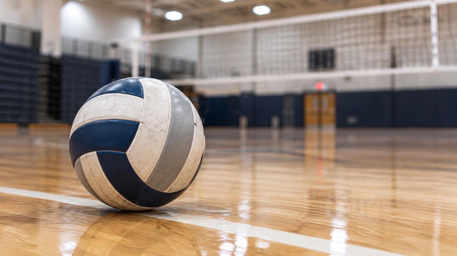 A volleyball resting on a hardwood gym floor near a net.