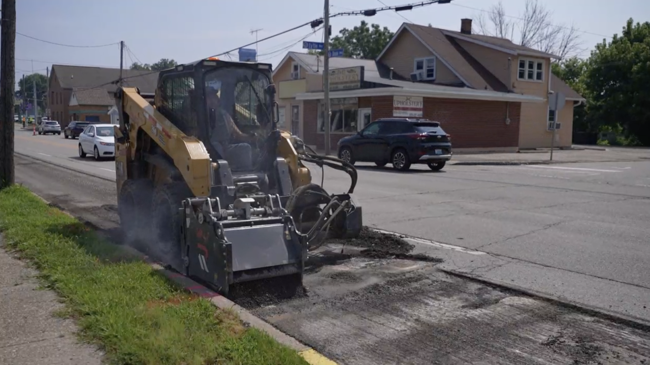 Yellow skid-steer milling machine grinding asphalt at a suburban road intersection.