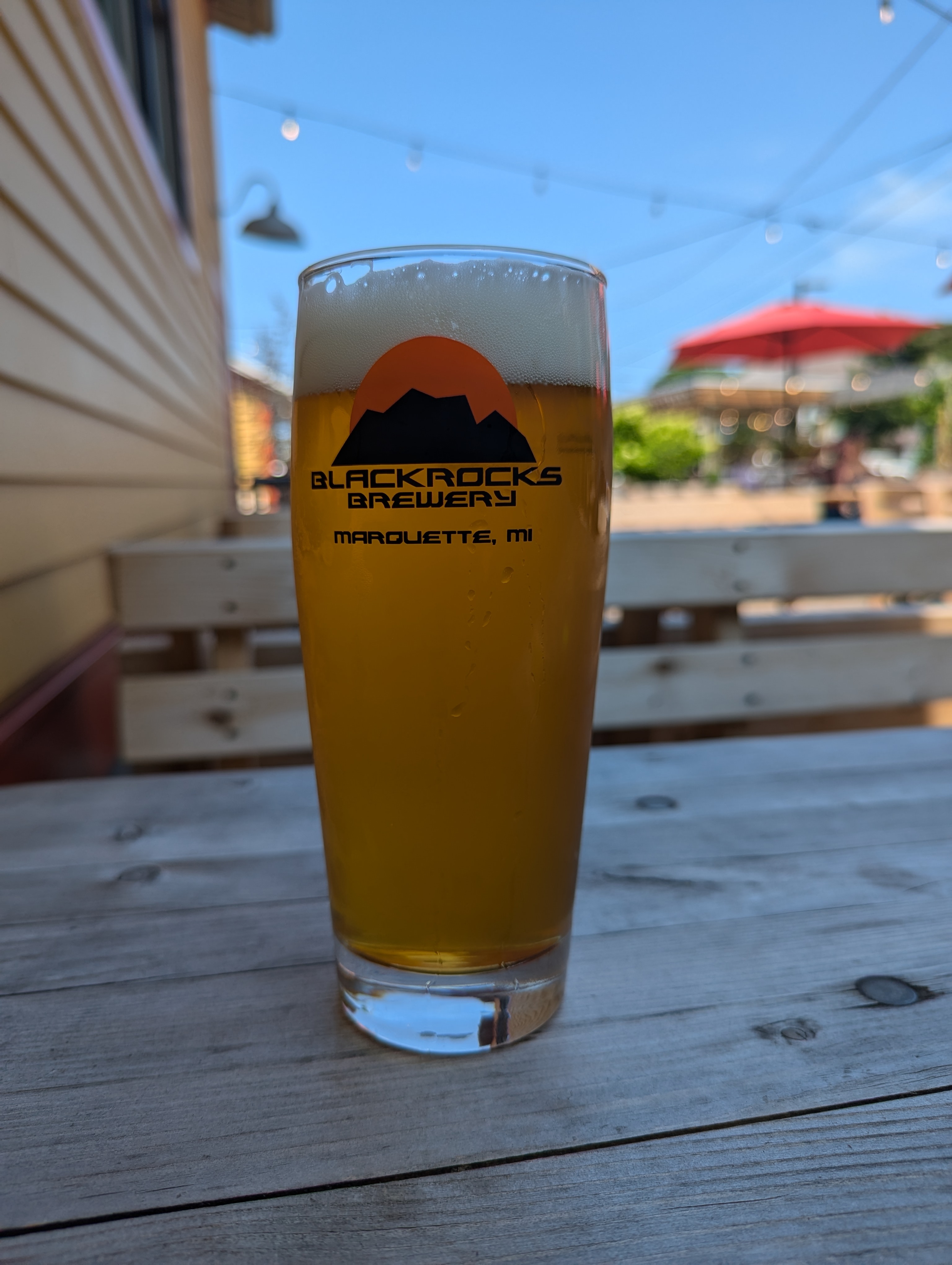 A branded pint glass of golden beer on an outdoor wooden table with string lights overhead.