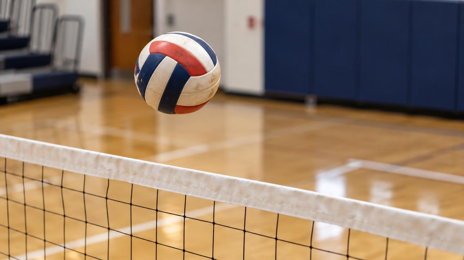 A volleyball suspended in mid-air above a net on a hardwood gymnasium court.