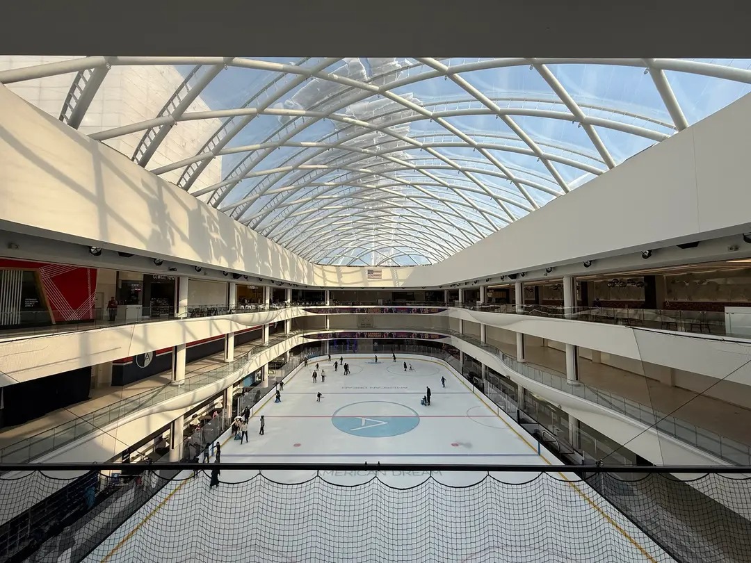 Aerial view of an indoor ice rink inside a multi-level shopping mall under a curved glass roof.