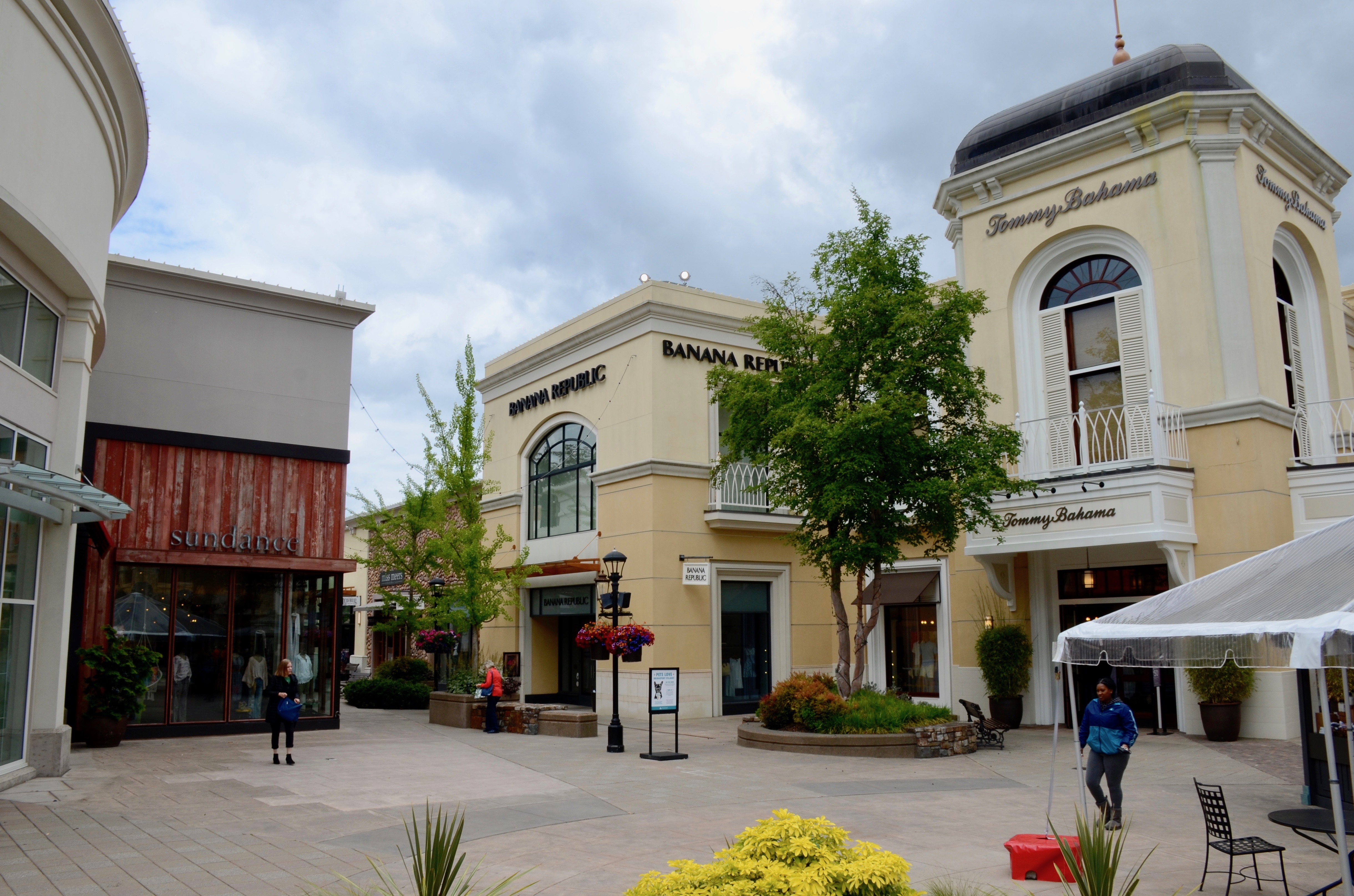 Outdoor shopping mall courtyard with retail storefronts, seating areas, and a white umbrella canopy.