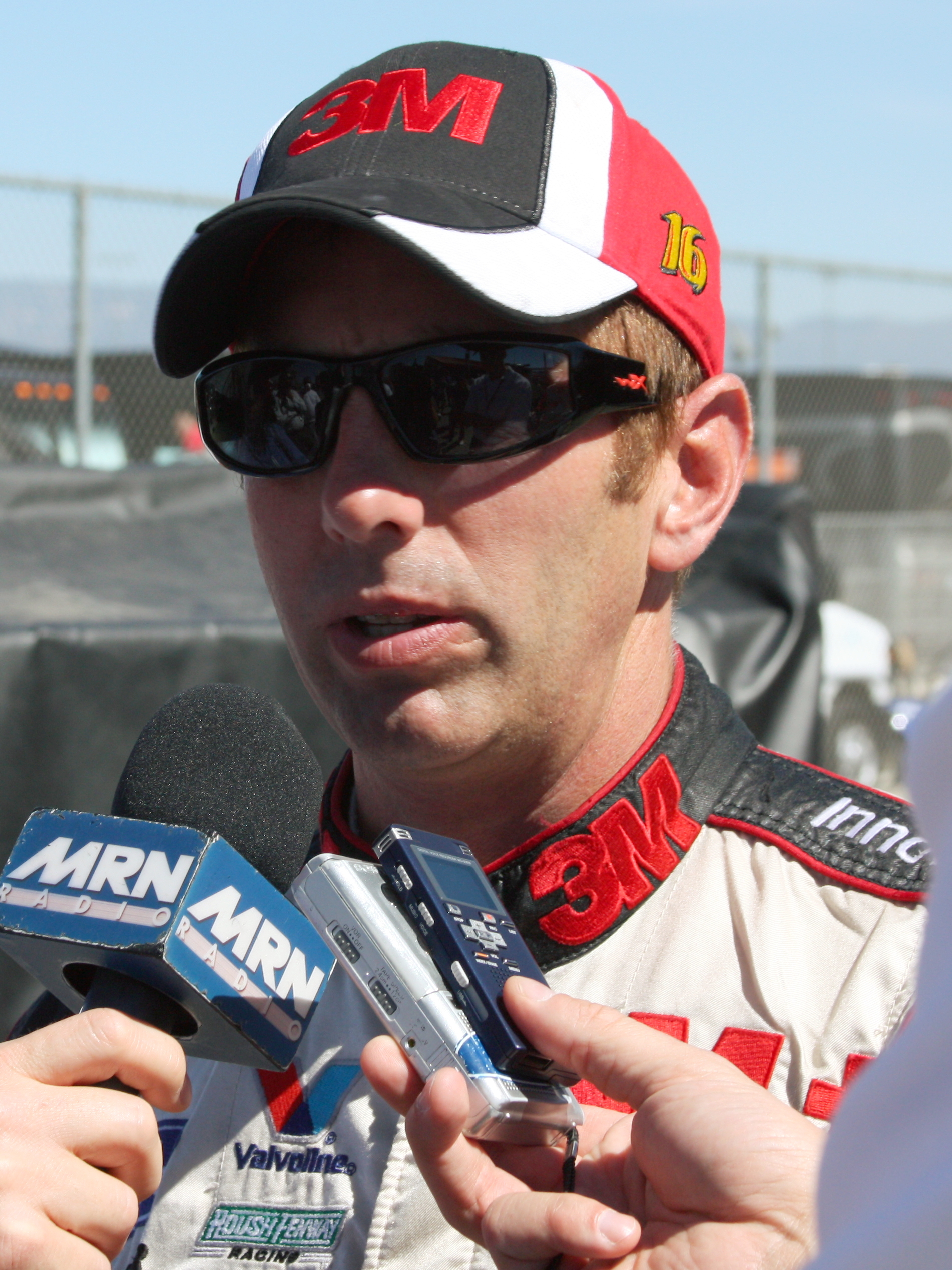 NASCAR driver in a 3M-sponsored racing suit speaking into multiple microphones at a race venue.