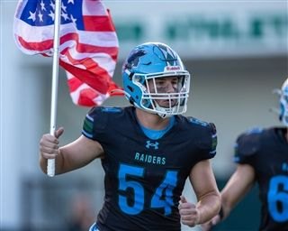 Football player in teal Raiders uniform carrying an American flag on the field.
