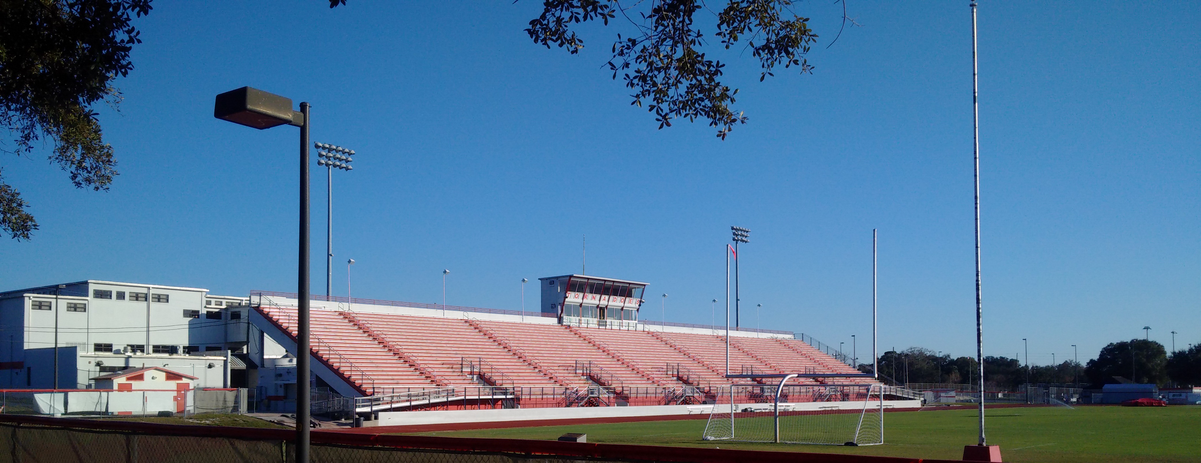 Outdoor stadium with tiered red bleachers, a press box, and tall light towers.