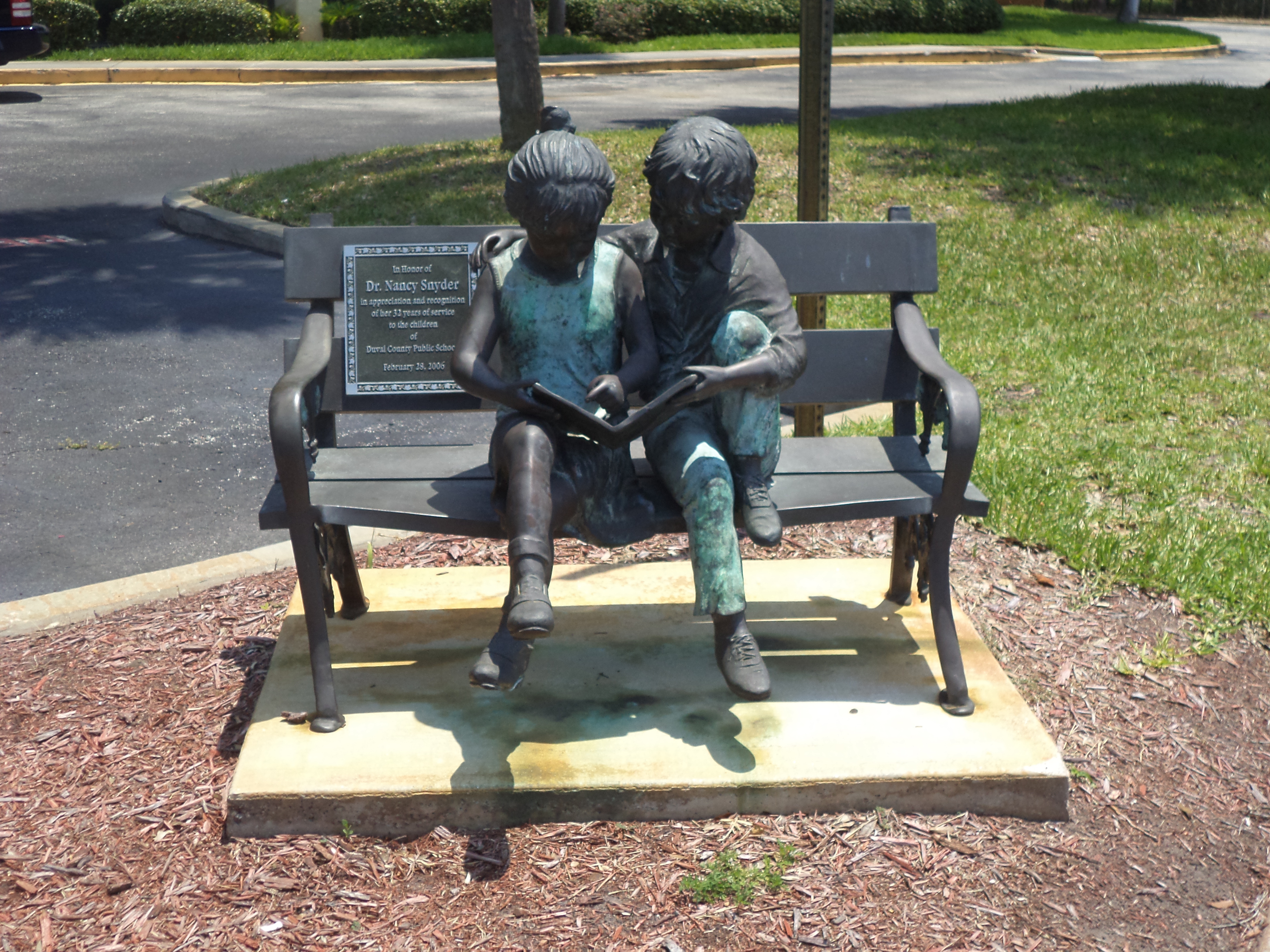 Bronze sculpture of two children seated on a bench sharing a book outside a school building.