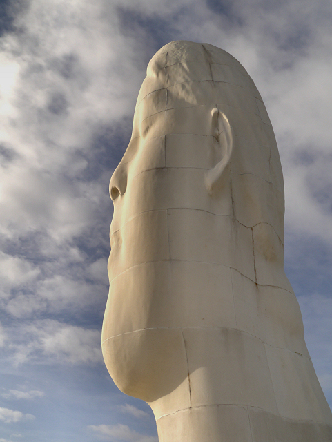 Large white sculptural head tilted skyward, photographed from below against a cloudy sky.